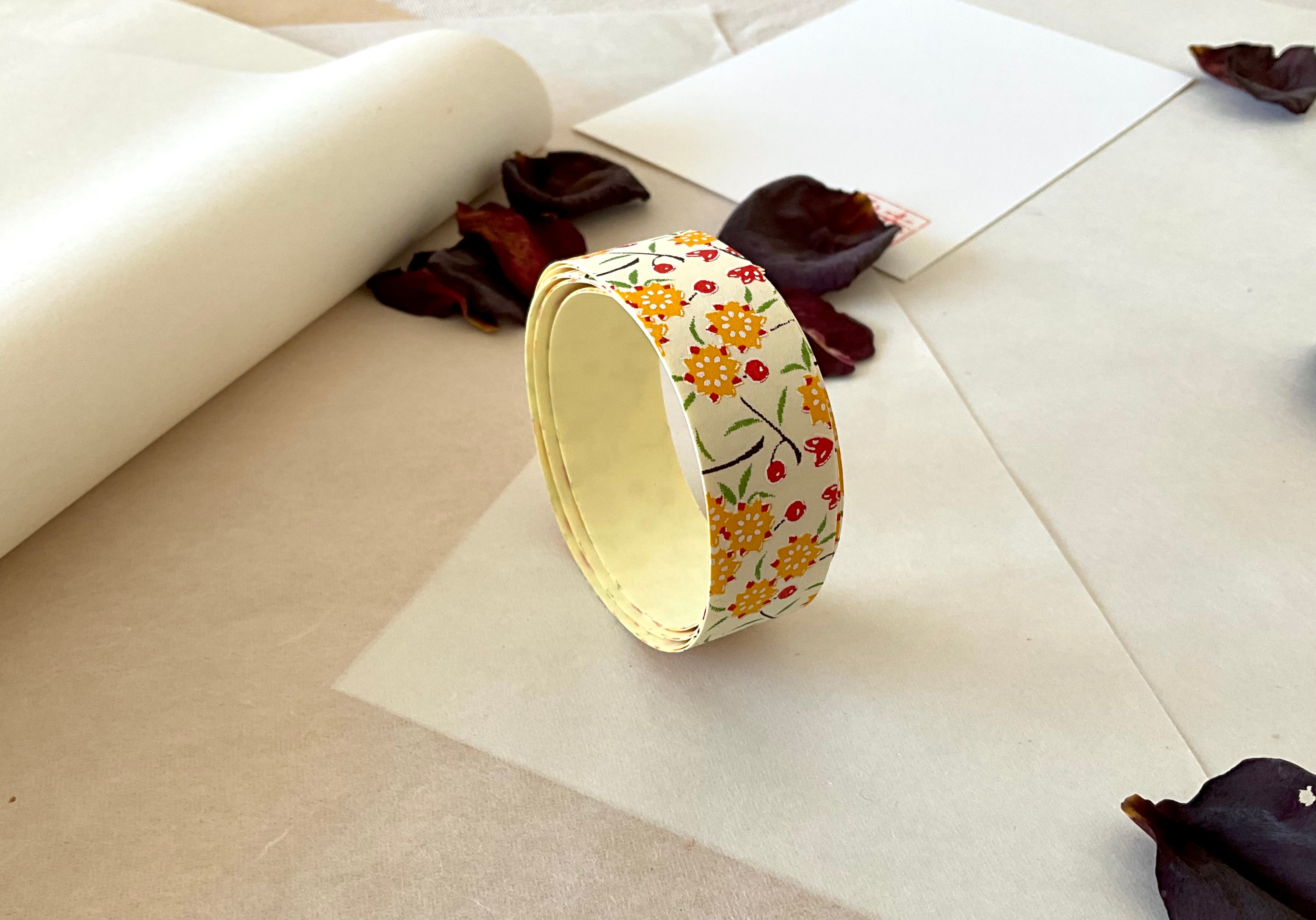 Roll of Shogado hand dyed yuzen washi tape standing upright on layered washi and kraft paper, Cream with yellow and red floral sprigs, cream adhesive backing visible, dried rose petals and hanko stamp card in background