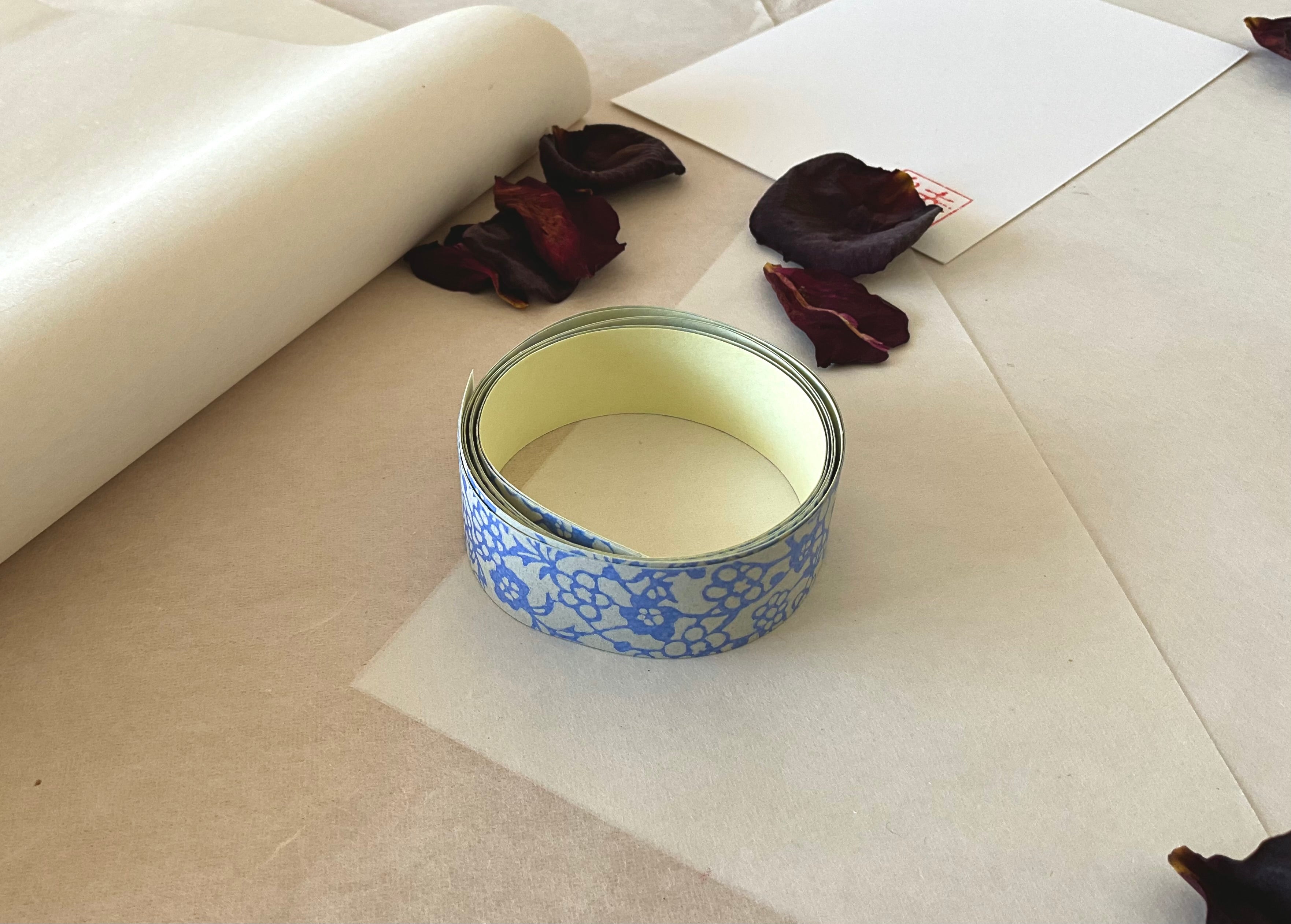Shogado yuzen washi tape unrolled on washi paper showing Grey-blue with blue floral block print pattern along its length, adhesive backing visible, dried rose petals nearby