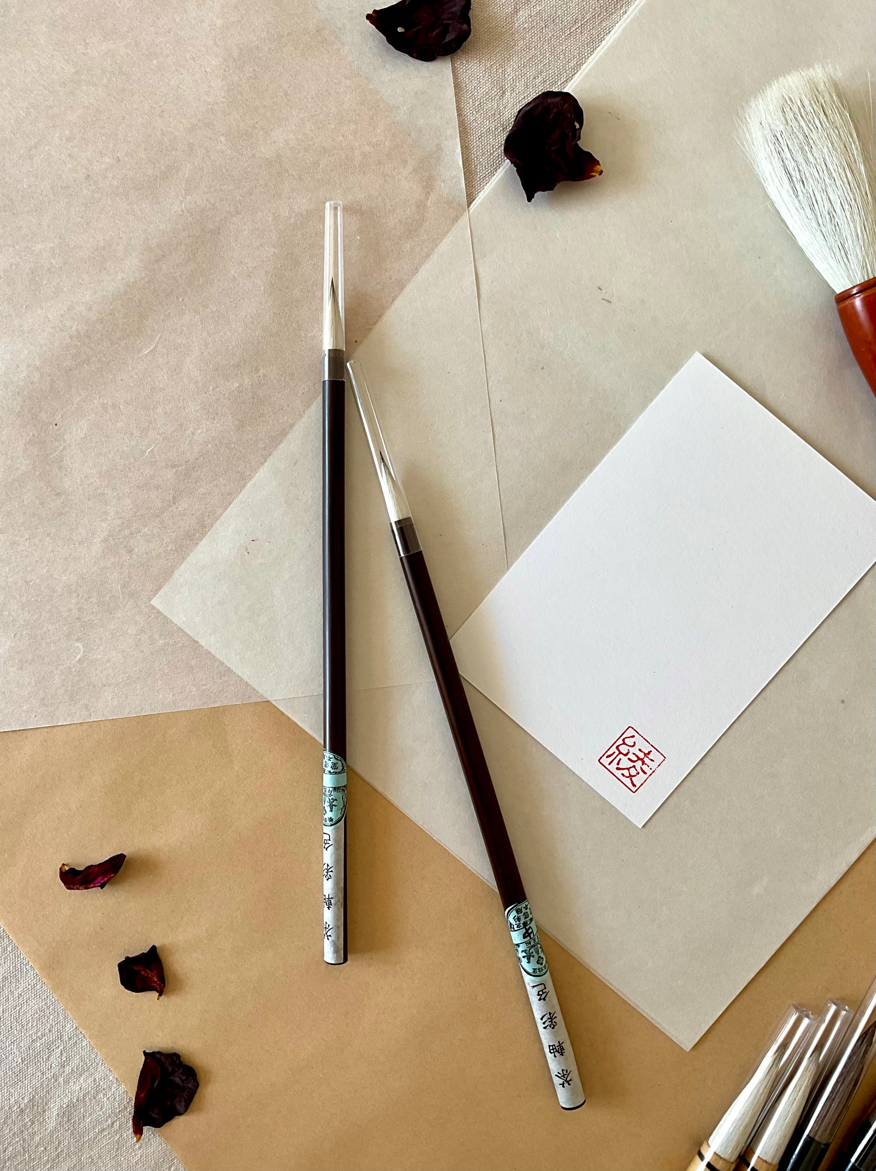Two Saiundo calligraphy brushes with dark lacquered handle and printed paper label, full pointed bristle, resting on layered washi paper with dried rose petals and a red hanko stamp card
