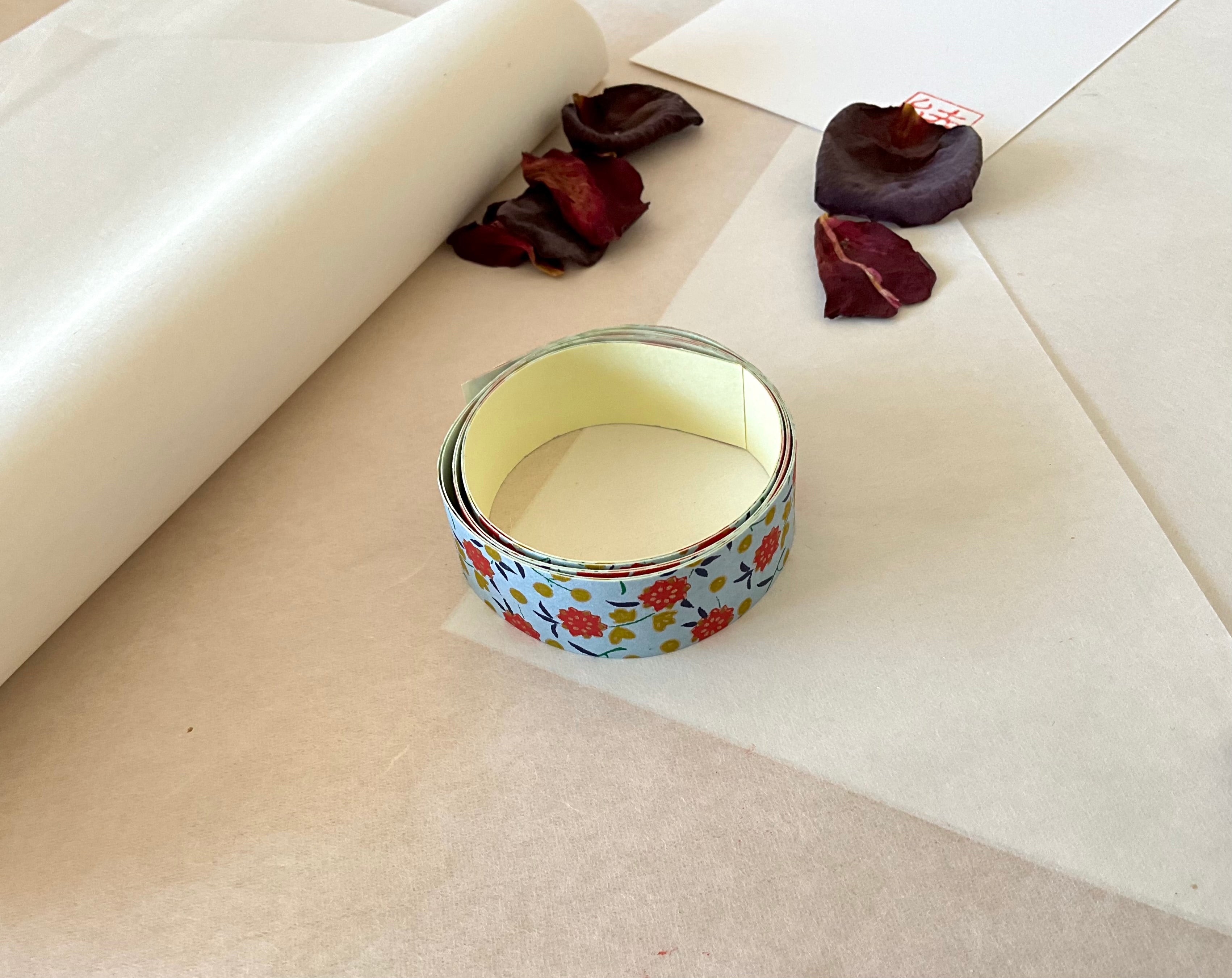Shogado yuzen washi tape unrolled on washi paper showing Pale blue with red flowers, mustard dots, navy leaves along its length, adhesive backing visible, dried rose petals nearby