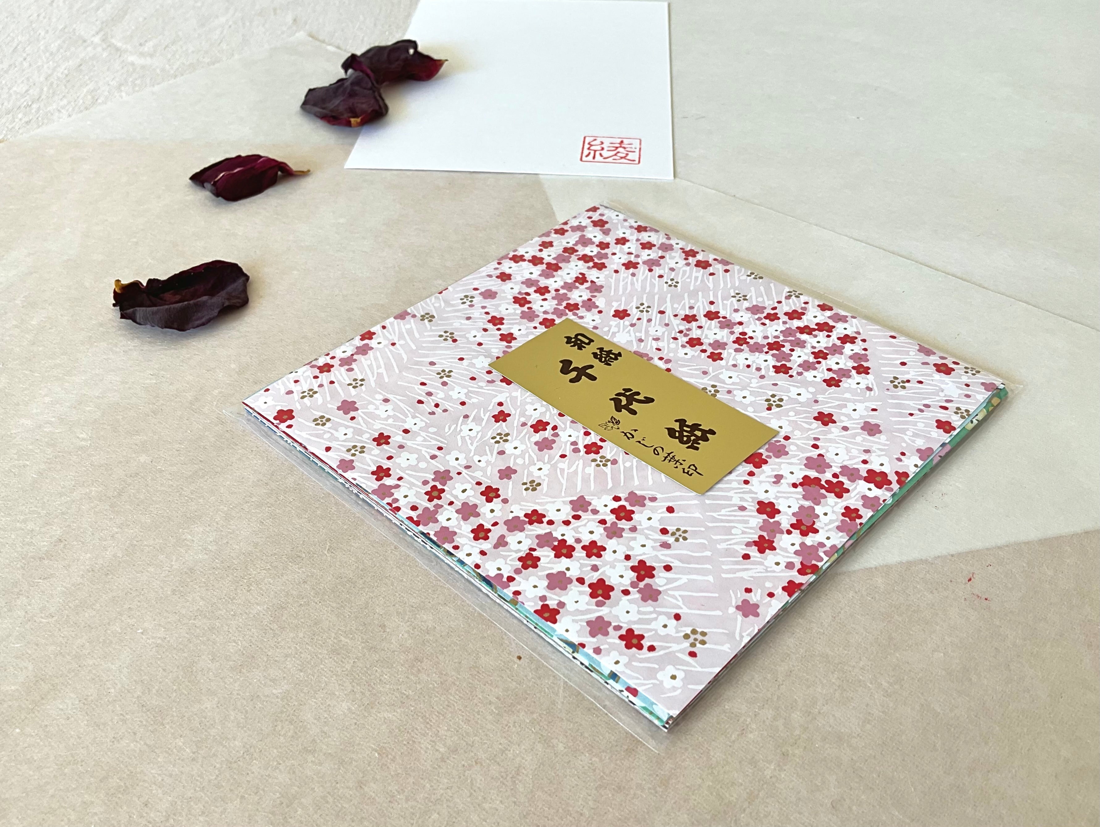 Morita Washi white and pink plum blossom chiyogami origami paper pack at an angle showing 20 sheets inside clear sleeve, on layered washi paper with dried rose petals