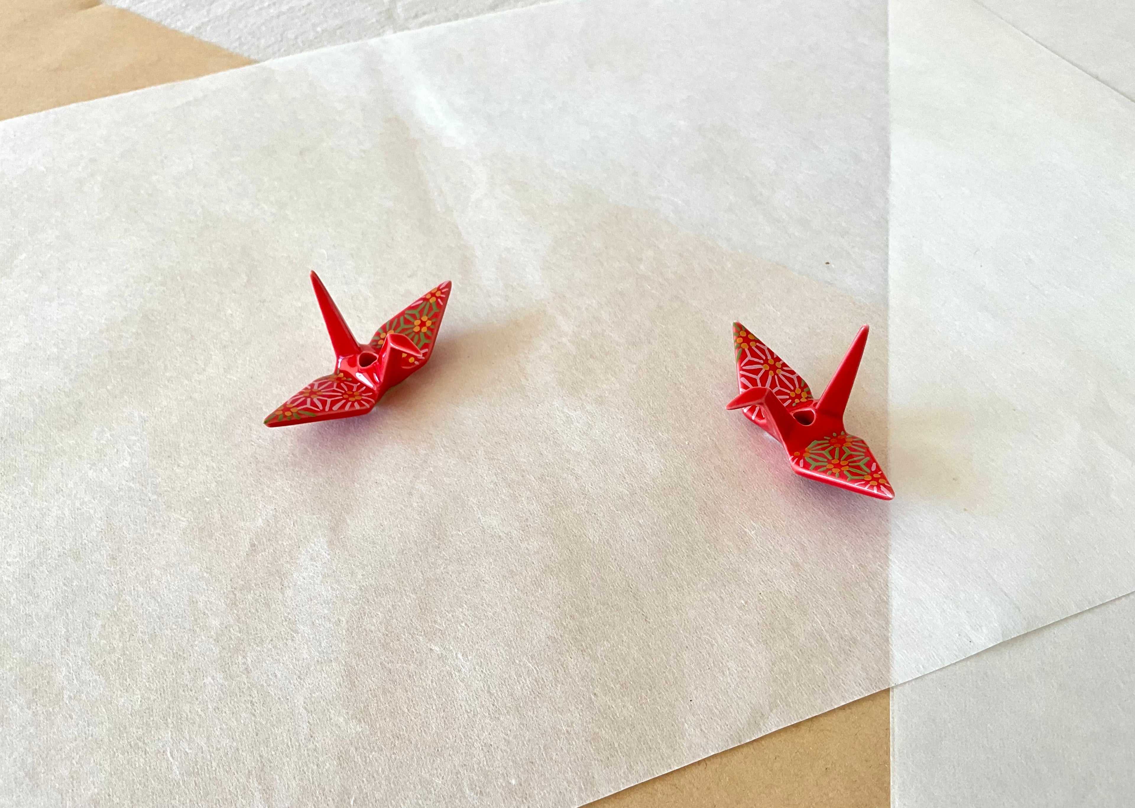 Two ceramic origami crane incense holders on washi paper, glazed in a red chiyogami pattern with gold and green floral motifs, each with a small hole at the back for an incense stick