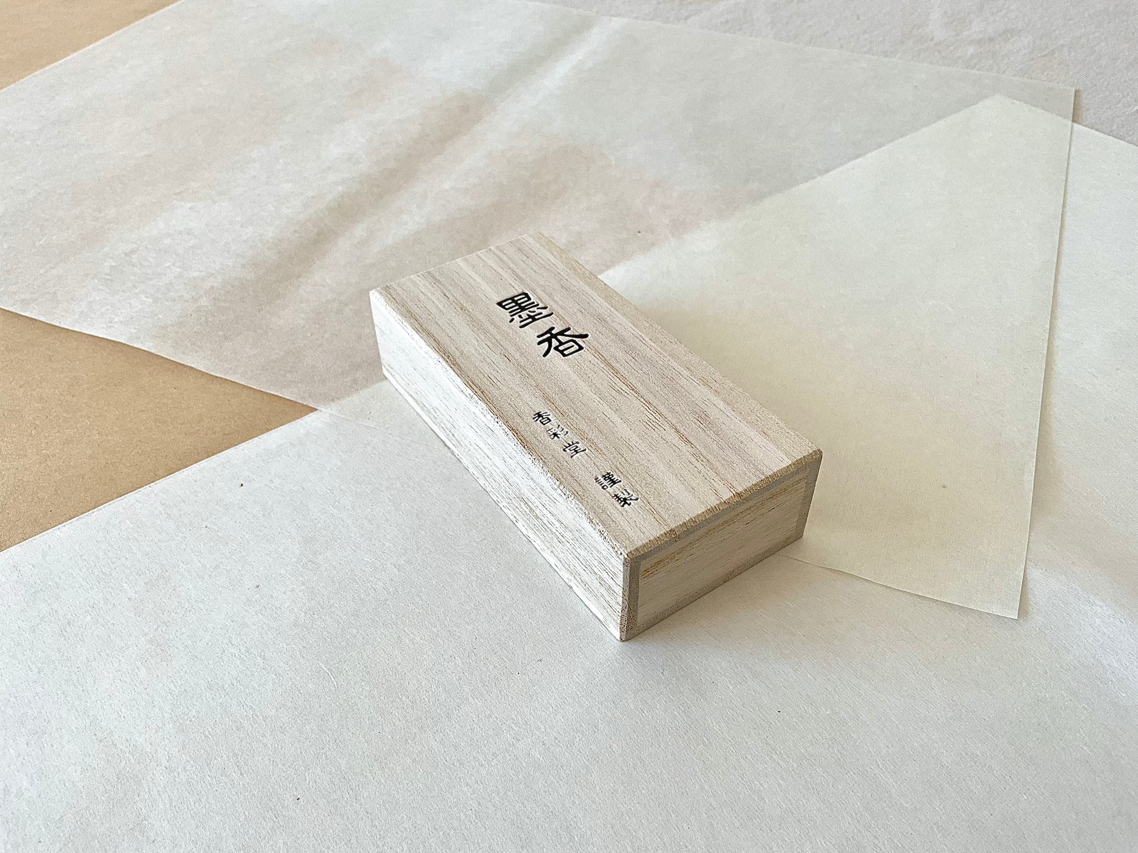 Kousaido Boku-ko Ink Fragrance paulownia wood box at an angle on layered washi paper, natural pale wood grain with Japanese ink fragrance kanji, Kousaido maker mark, clean minimal composition