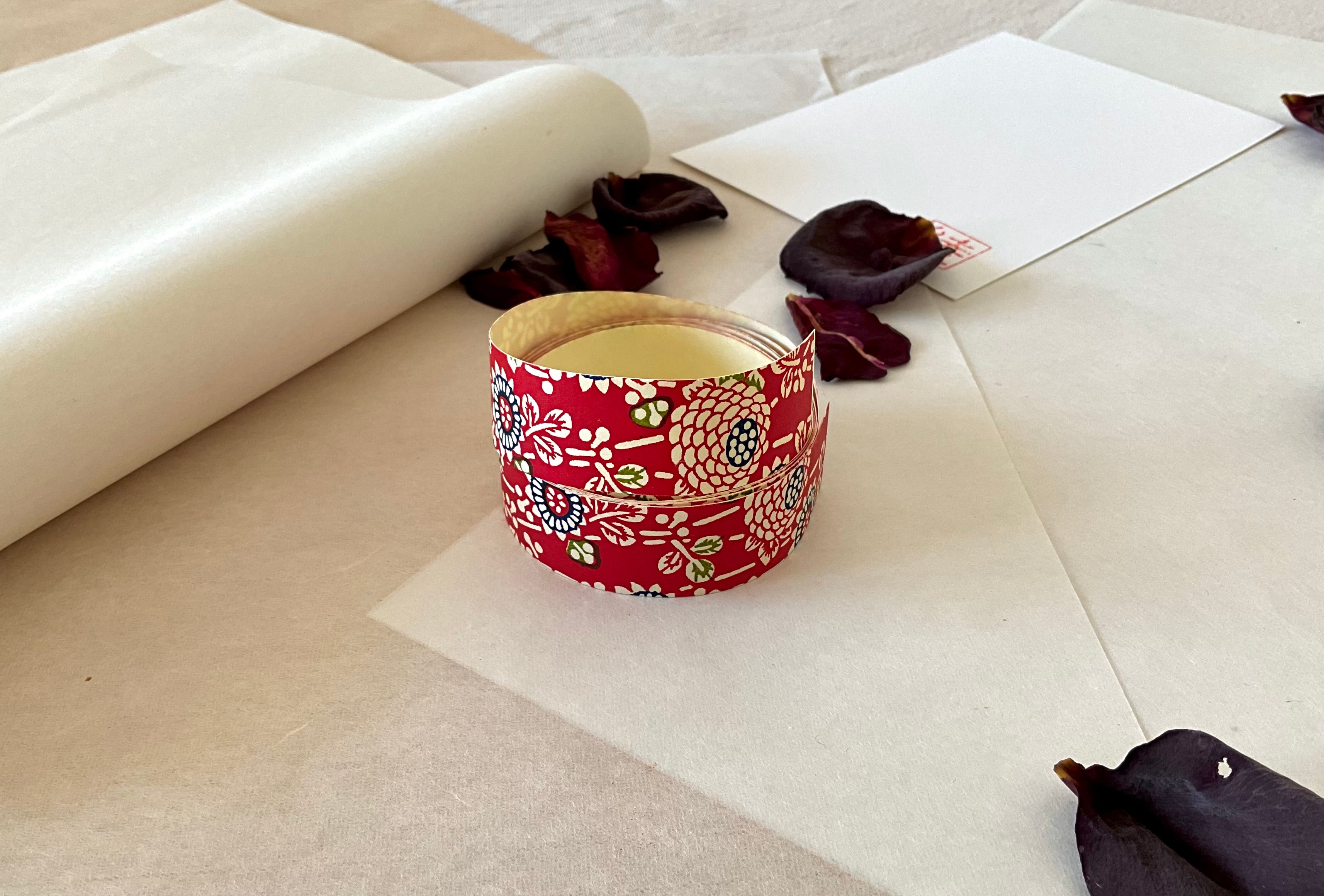 Both sheets of Shogado hand dyed yuzen washi tape laid side by side on washi paper, showing Crimson red with cream chrysanthemum and floral motifs, navy dotted circle centers, green accents repeated across both strips, dried rose petals nearby