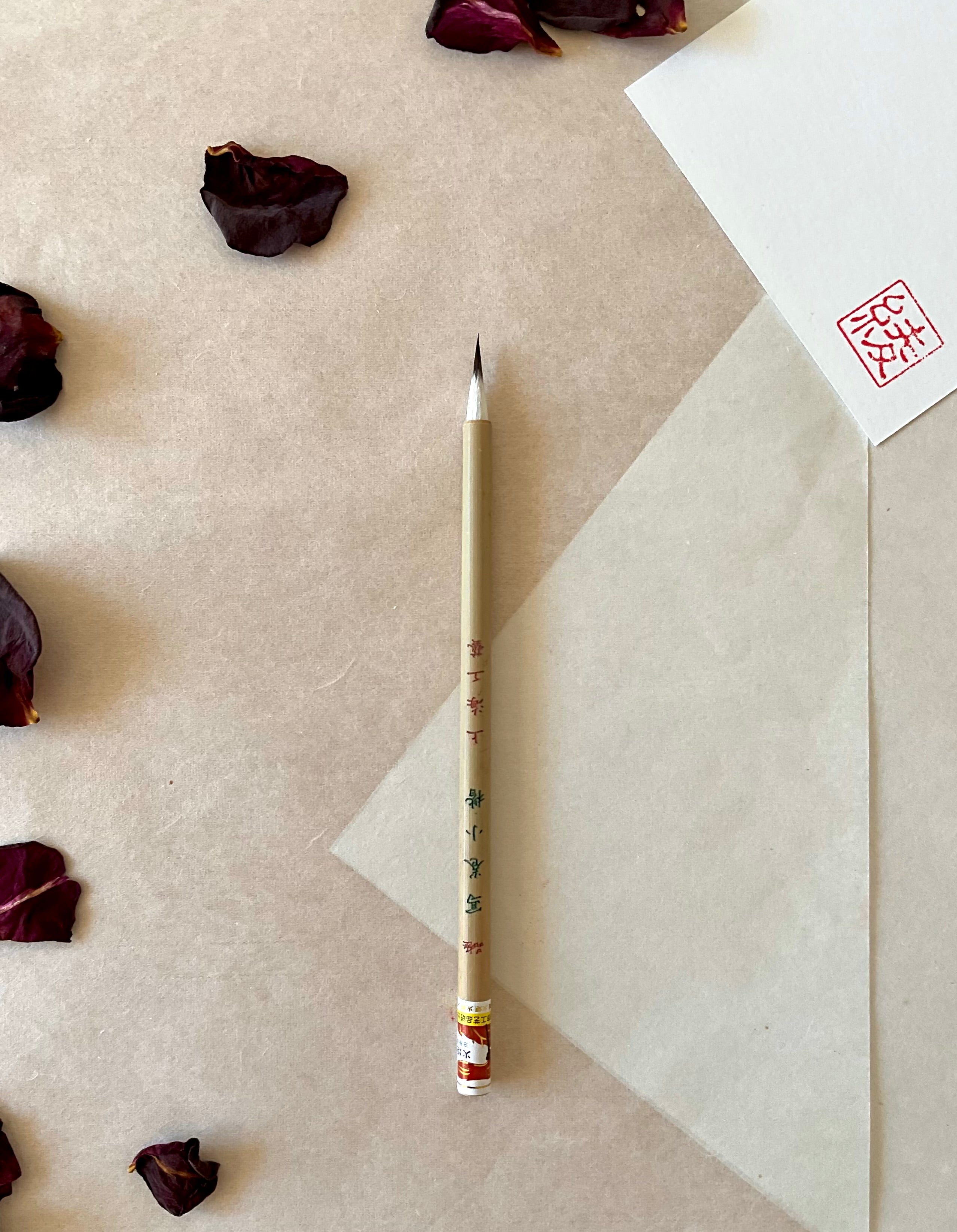 Kobaien fine detail calligraphy brush with sharp pointed tip and bamboo handle printed with Japanese maker markings, resting on layered washi paper with dried rose petals and a red hanko stamp card