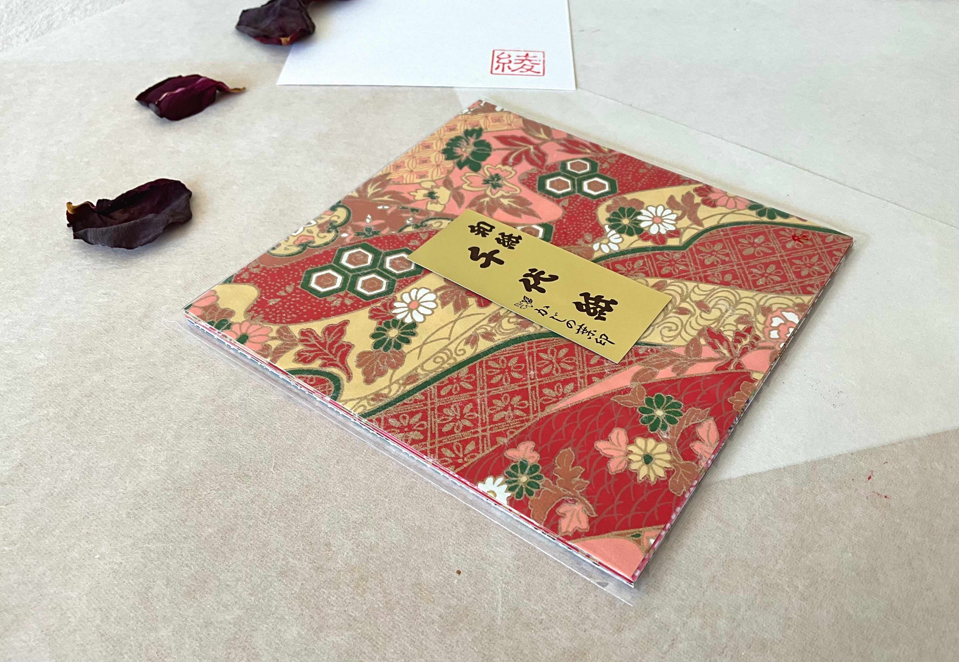 Morita Washi mixed floral patchwork chiyogami paper pack at an angle showing layered sheets inside sleeve, rich red and gold kimono-inspired cover, on washi paper with dried rose petals