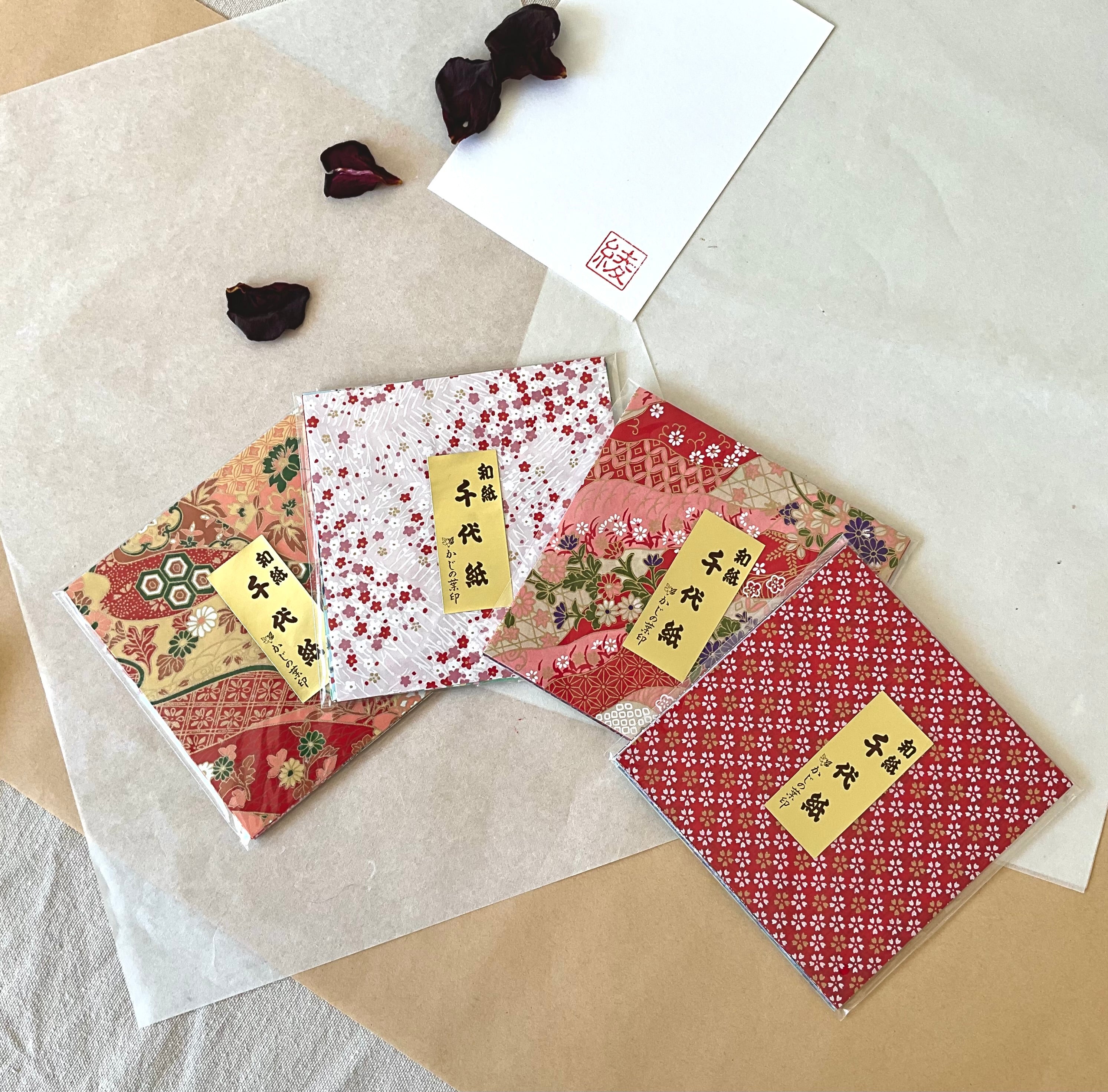 Four packs of Morita Washi chiyogami origami paper fanned out on washi and kraft paper, showing four different traditional Japanese pattern covers in reds, pink and gold, with dried rose petals and hanko stamp card