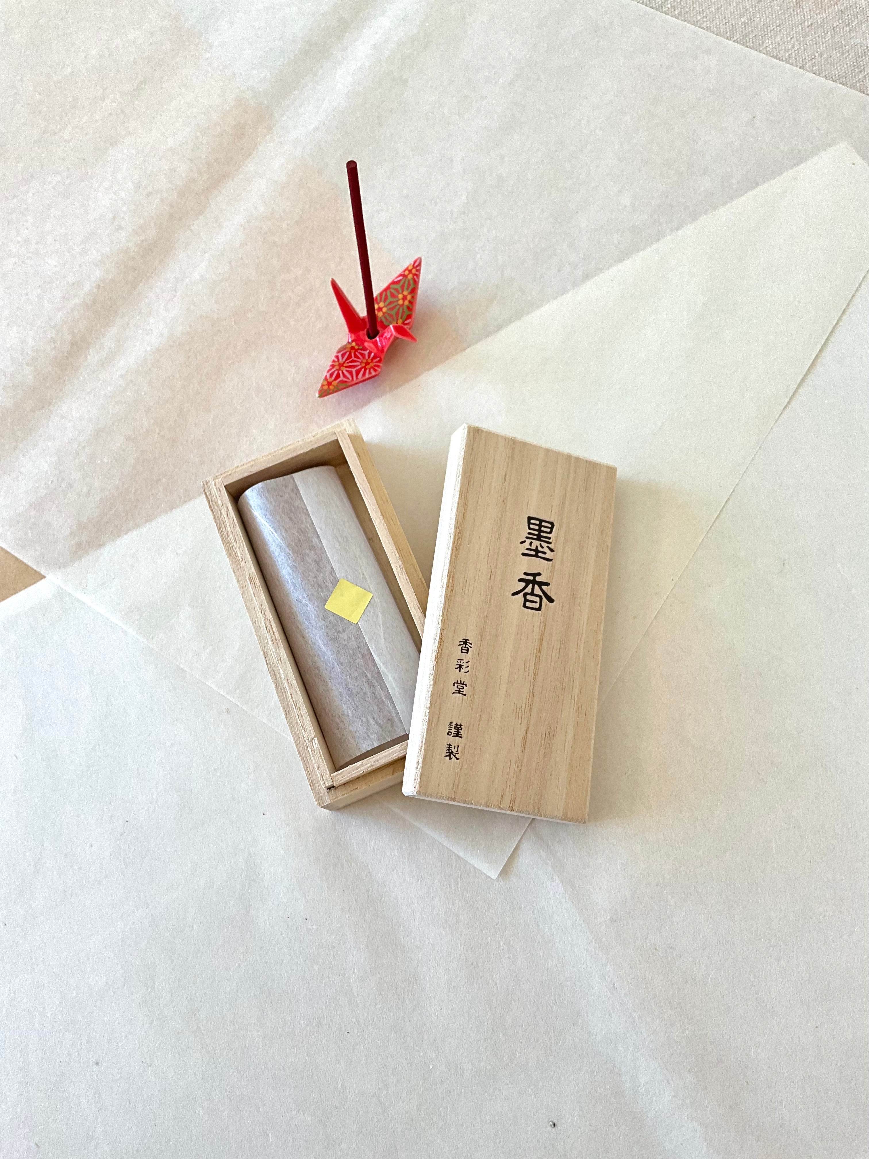 Kousaido Boku-ko Ink Fragrance incense paulownia wood box open showing incense sticks in tissue, lid with kanji beside it, small red chiyogami origami crane incense holder with lit stick on layered washi paper