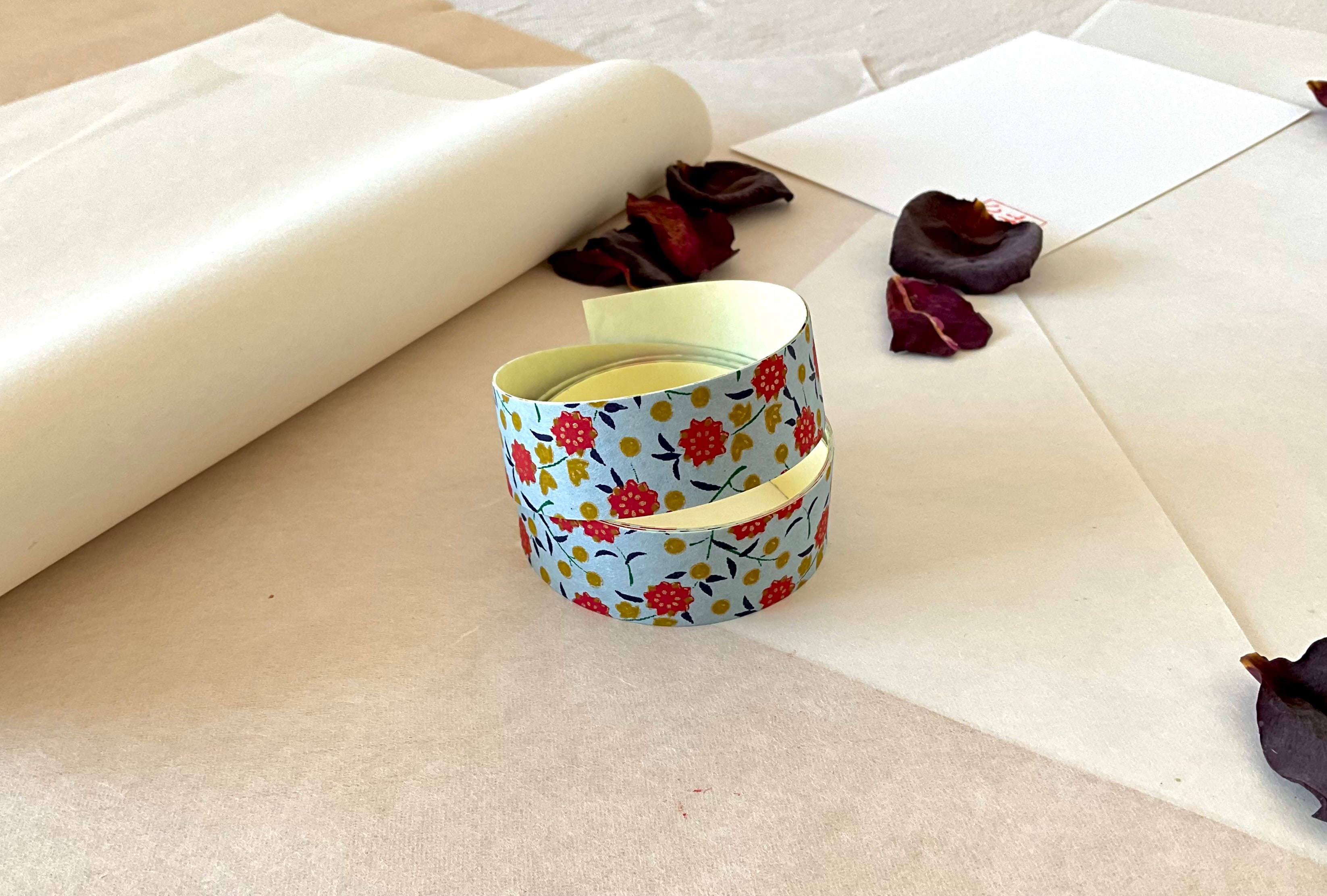 Both sheets of Shogado hand dyed yuzen washi tape laid on top of each other washi paper, showing Pale blue with red flowers, mustard dots, navy leaves repeated across both strips, dried rose petals nearby
