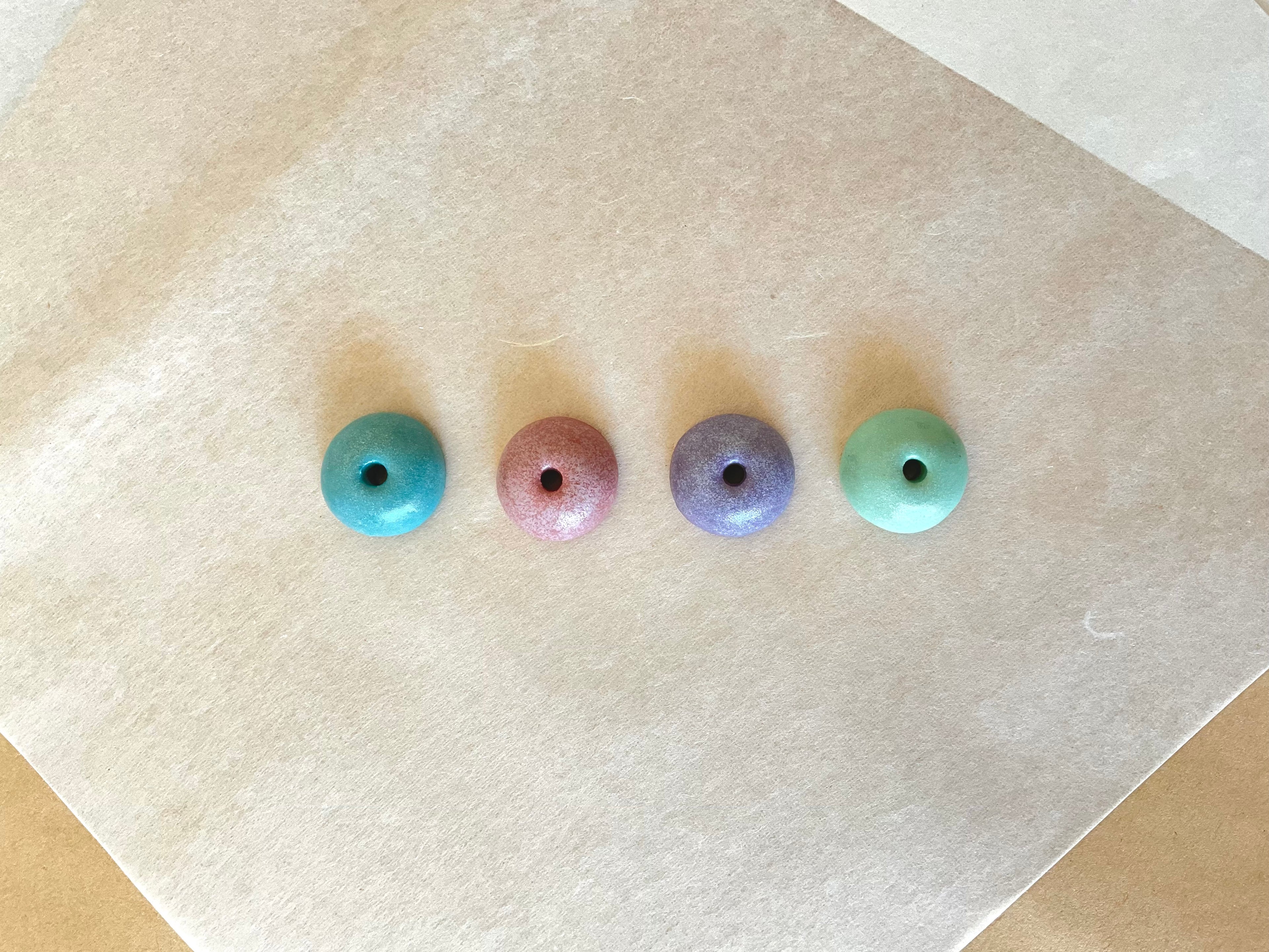 Four round ceramic incense holders in a row on washi paper, glazed in teal, dusty rose, lavender and sage green, each with a small central hole for an incense stick