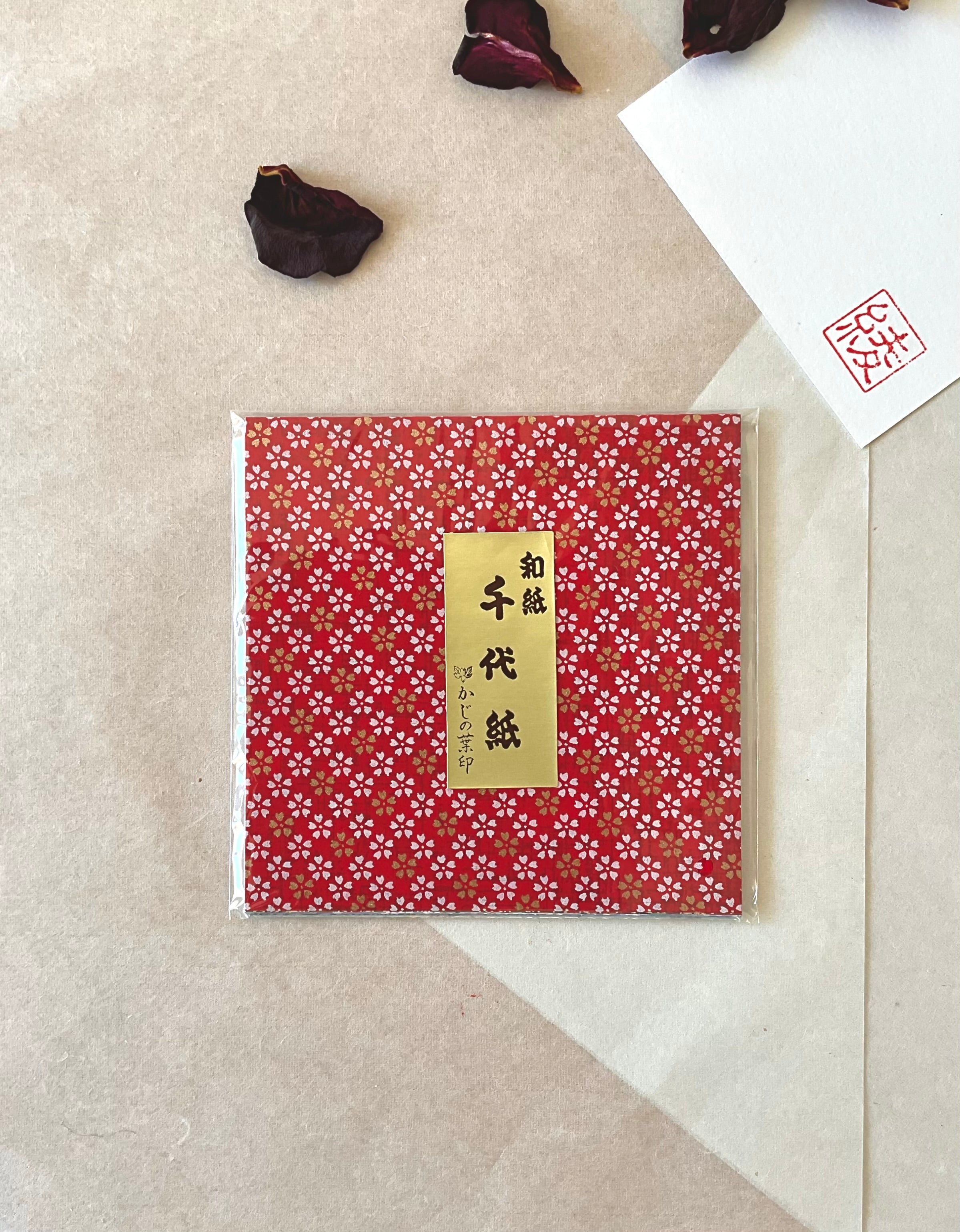 Single sealed pack of Morita Washi chiyogami origami paper with deep red and gold sakura repeat pattern, gold Japanese label, on neutral washi paper background with hanko stamp card