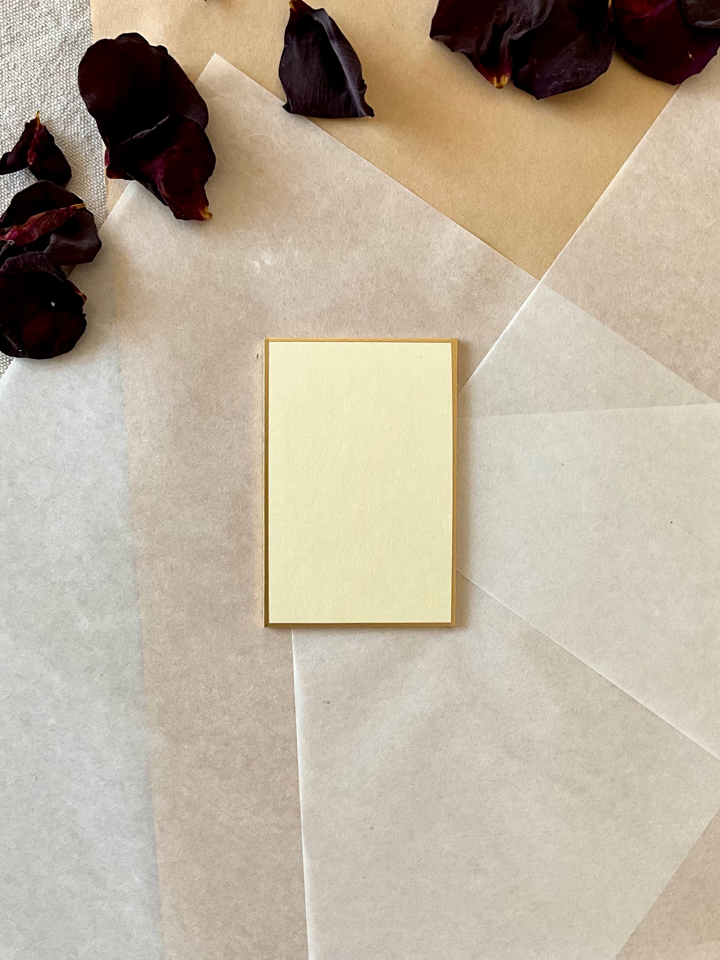 Single small rectangle cream Kyukyodo shikishi board centered on layered washi and kraft paper with dried dark rose petals at upper left, showing the clean square form and gold-painted edge