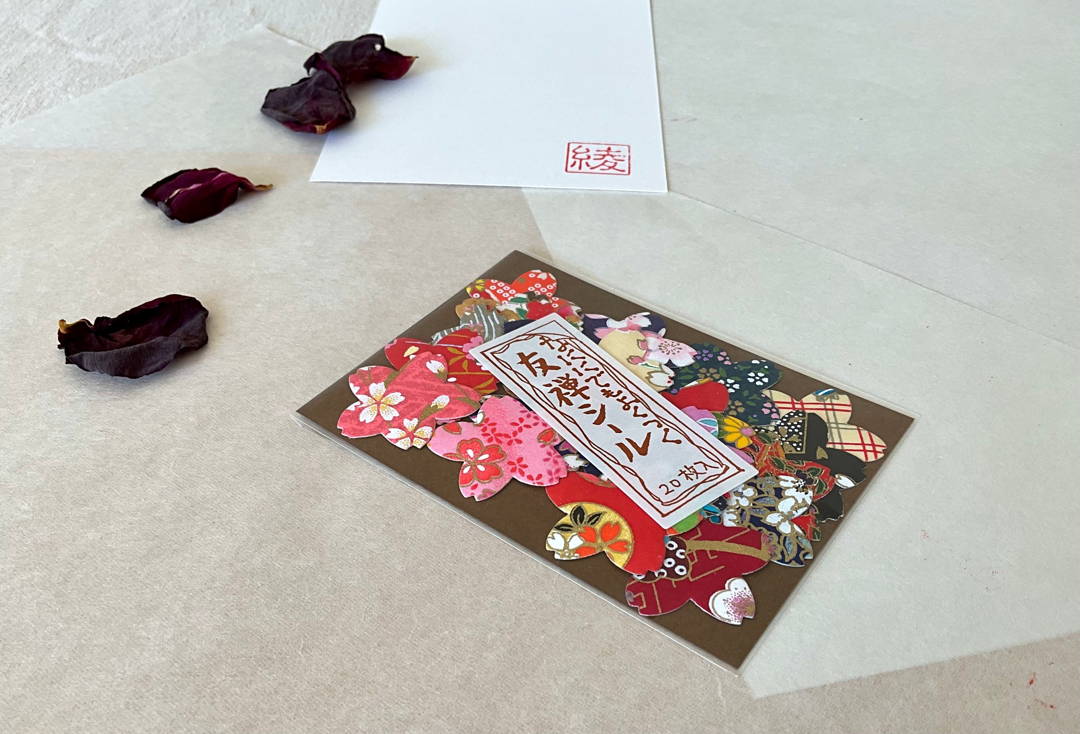 Morita Washi yuzen sticker pack at an angle on washi paper showing the layered edge of the pack and mixed sticker designs in red sakura, pink floral and dark patterned chiyogami shapes, with hanko stamp card in background