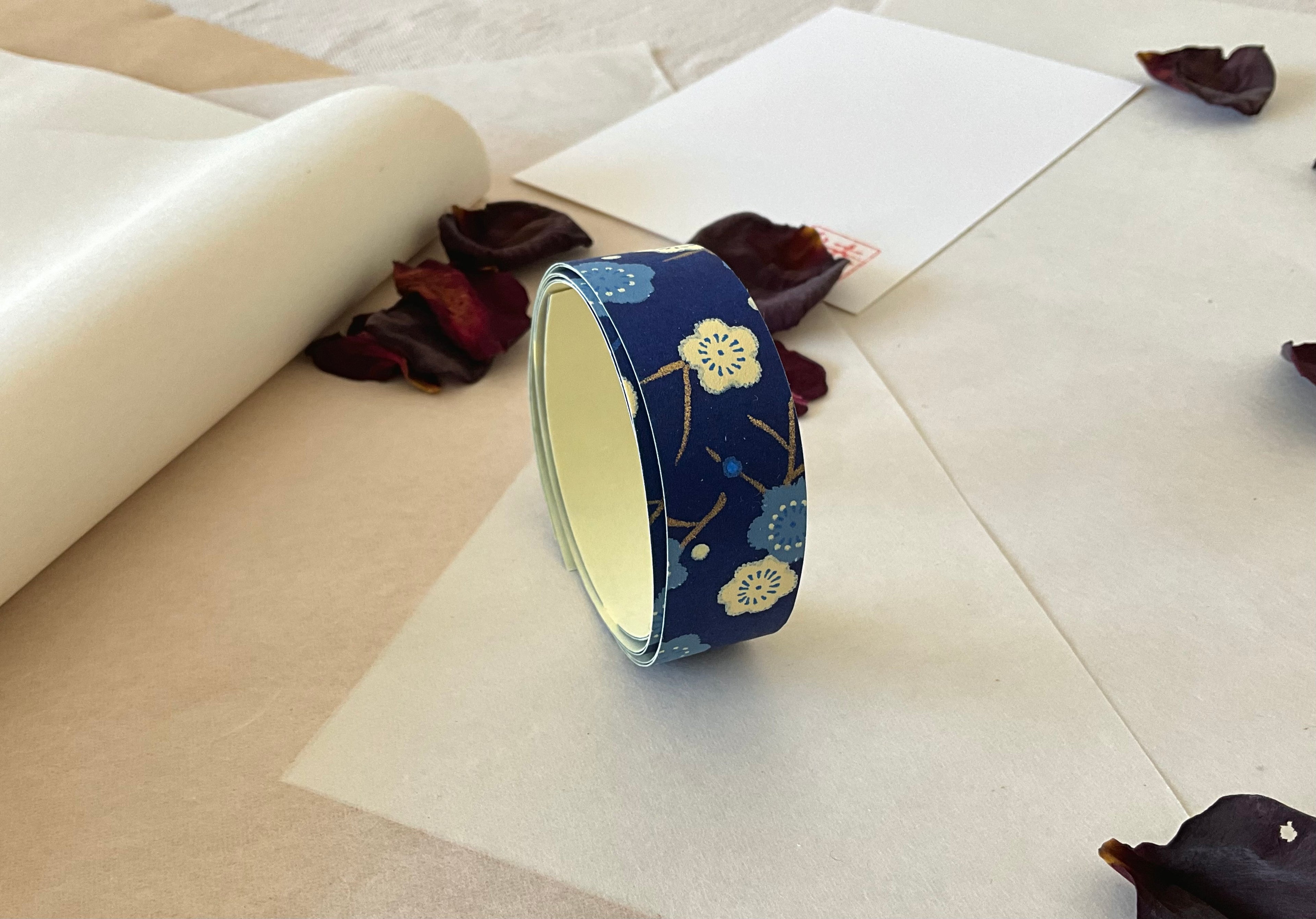 Roll of Shogado hand dyed yuzen washi tape standing upright on layered washi and kraft paper, Navy with cream and pale blue plum blossom branches, cream adhesive backing visible, dried rose petals and hanko stamp card in background