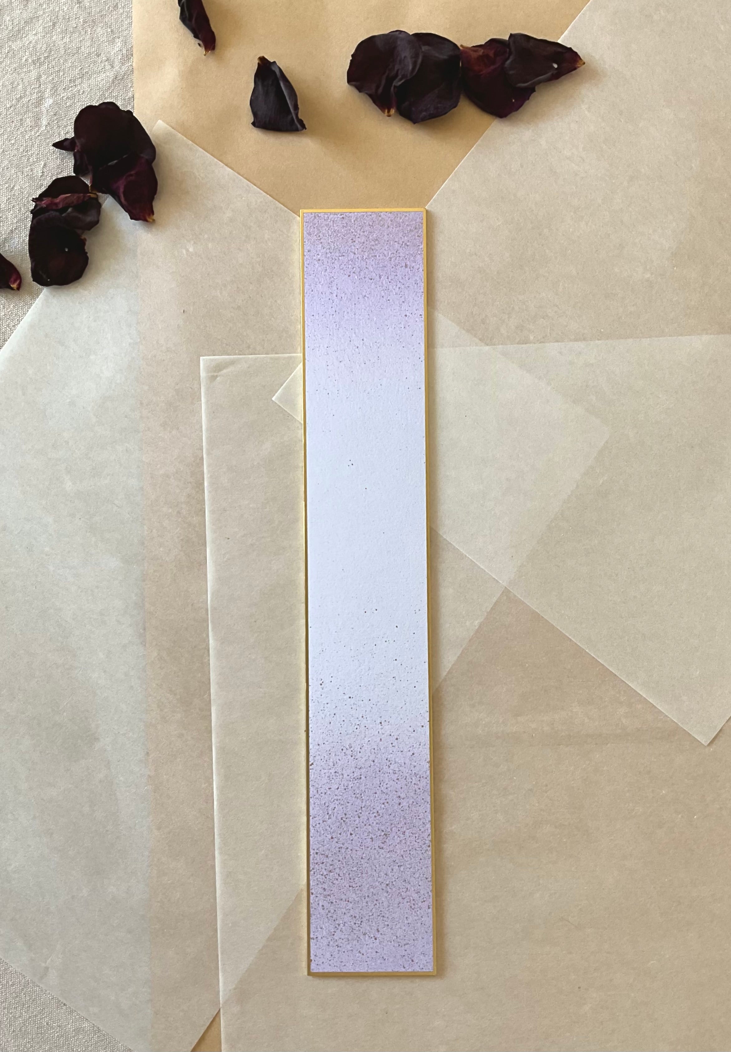 Single Kyukyodo tanzaku strip in lavender with speckled texture and gold border, centered on layered washi and kraft paper with scattered dried rose petals, top-down view