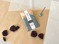 Single Kousaidou Arashiyama Moonlight incense pack on layered kraft and washi paper, green and gold illustrated sleeve with Japanese calligraphy title, lit dark incense stick standing in small celadon round holder, surrounded by dried rose petals