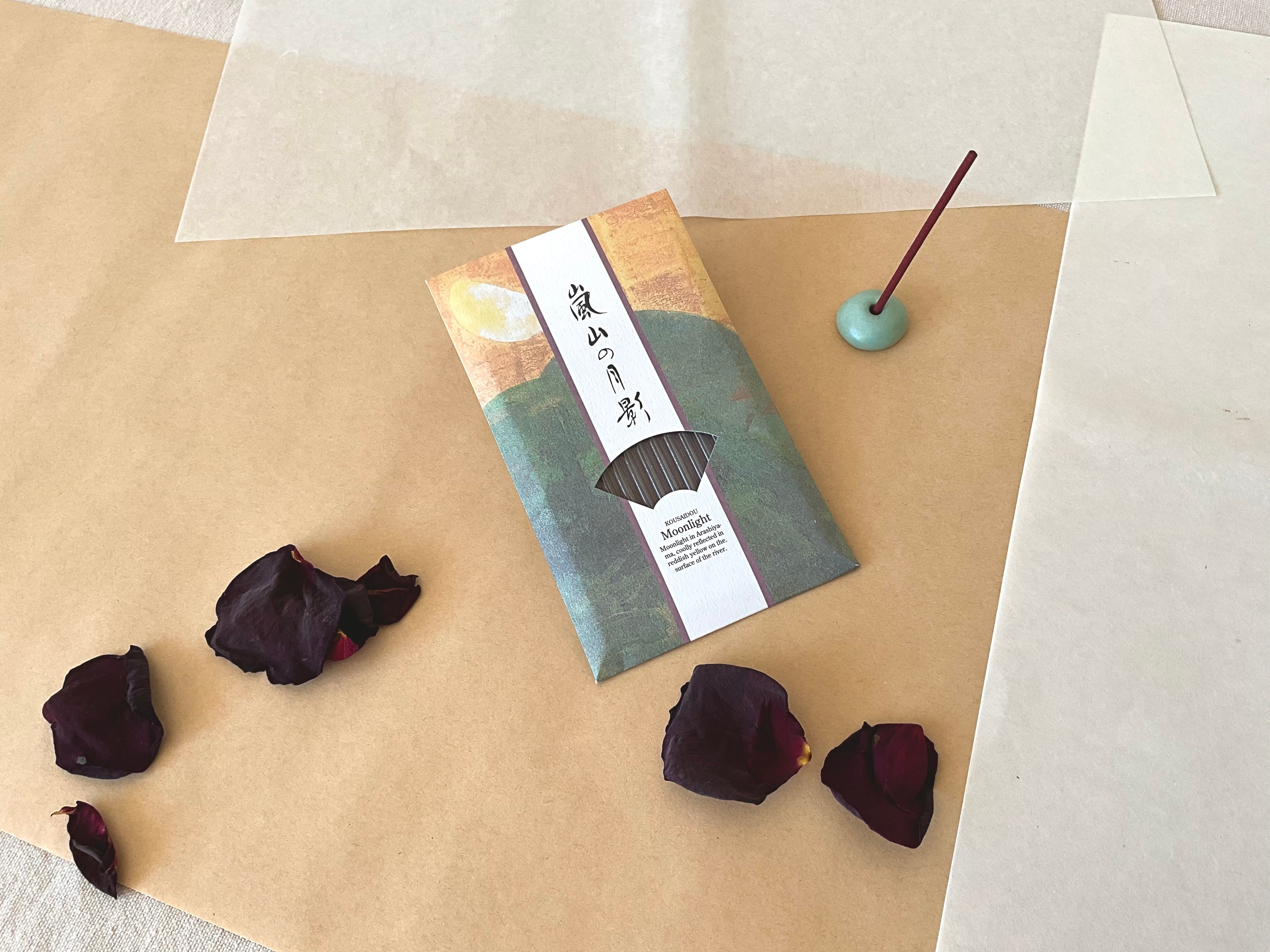 Single Kousaidou Arashiyama Moonlight incense pack on layered kraft and washi paper, green and gold illustrated sleeve with Japanese calligraphy title, lit dark incense stick standing in small celadon round holder, surrounded by dried rose petals