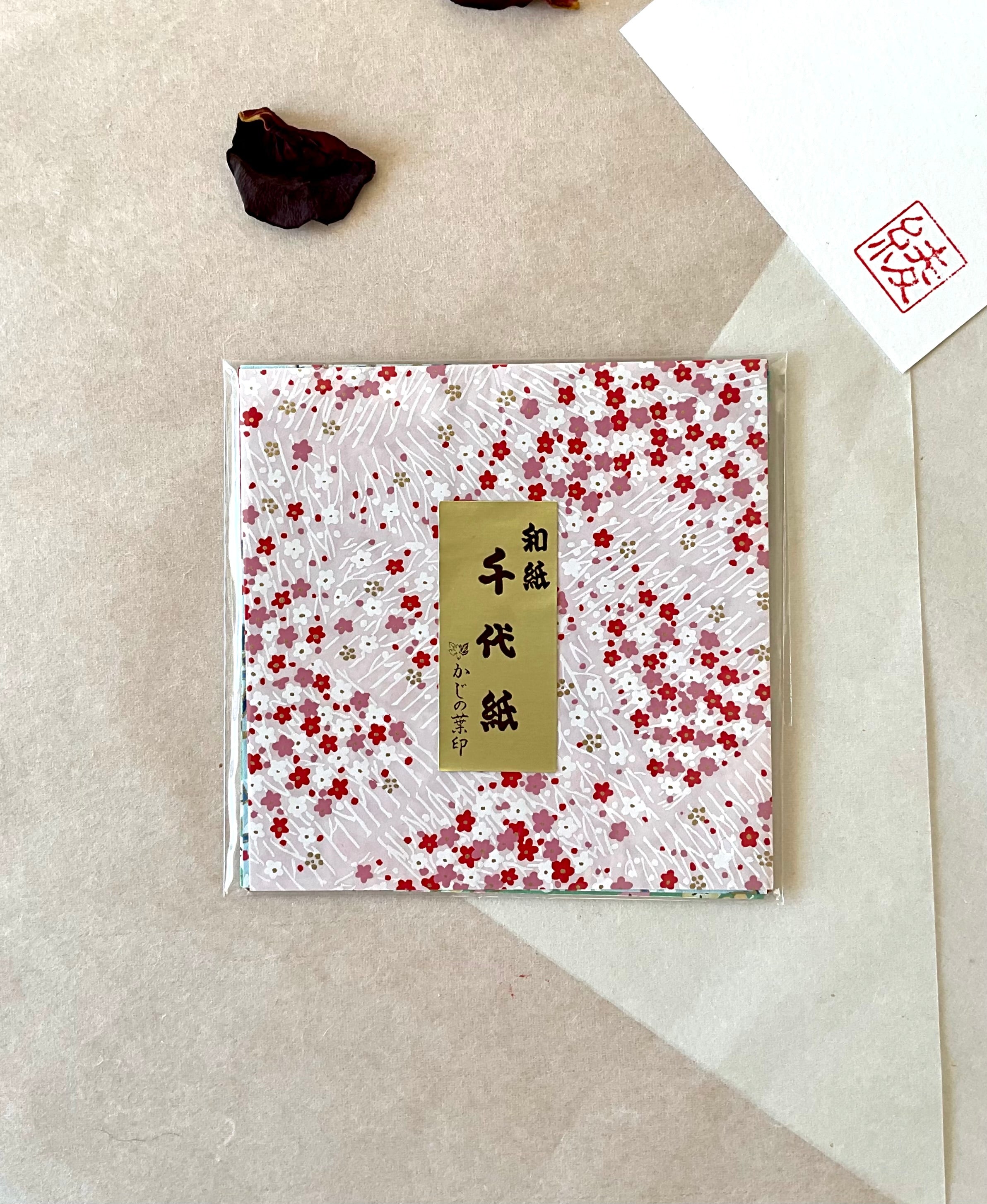 Single sealed pack of Morita Washi origami paper with white ground and scattered red, pink and mauve plum blossoms on bamboo branch pattern, gold label, on neutral washi background