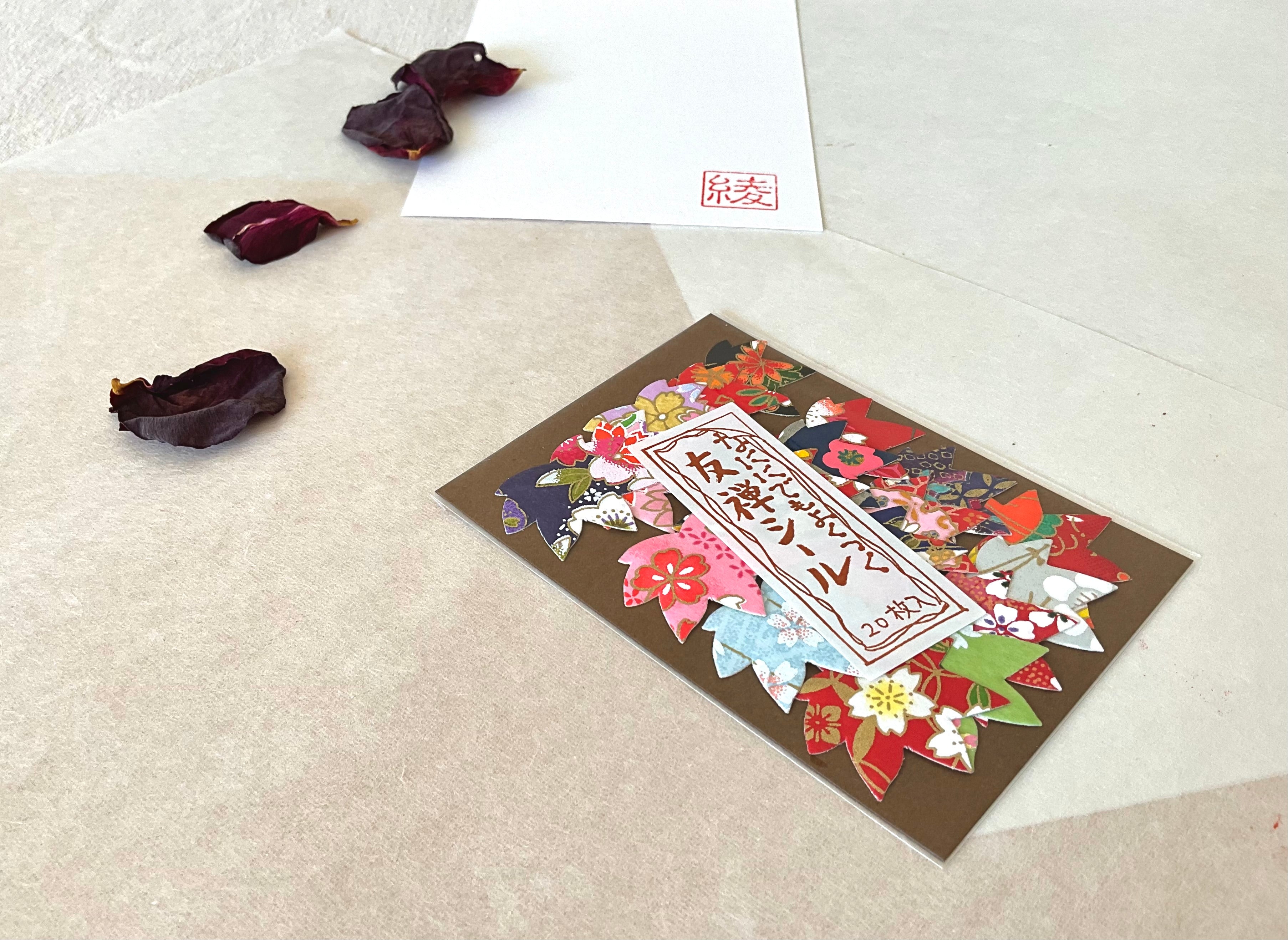 Morita Washi yuzen flower sticker pack photographed at a low angle on layered washi paper, showing the mixed sticker designs including light blue florals, red sakura and colorful petal shapes, with hanko stamp card and dried rose petals