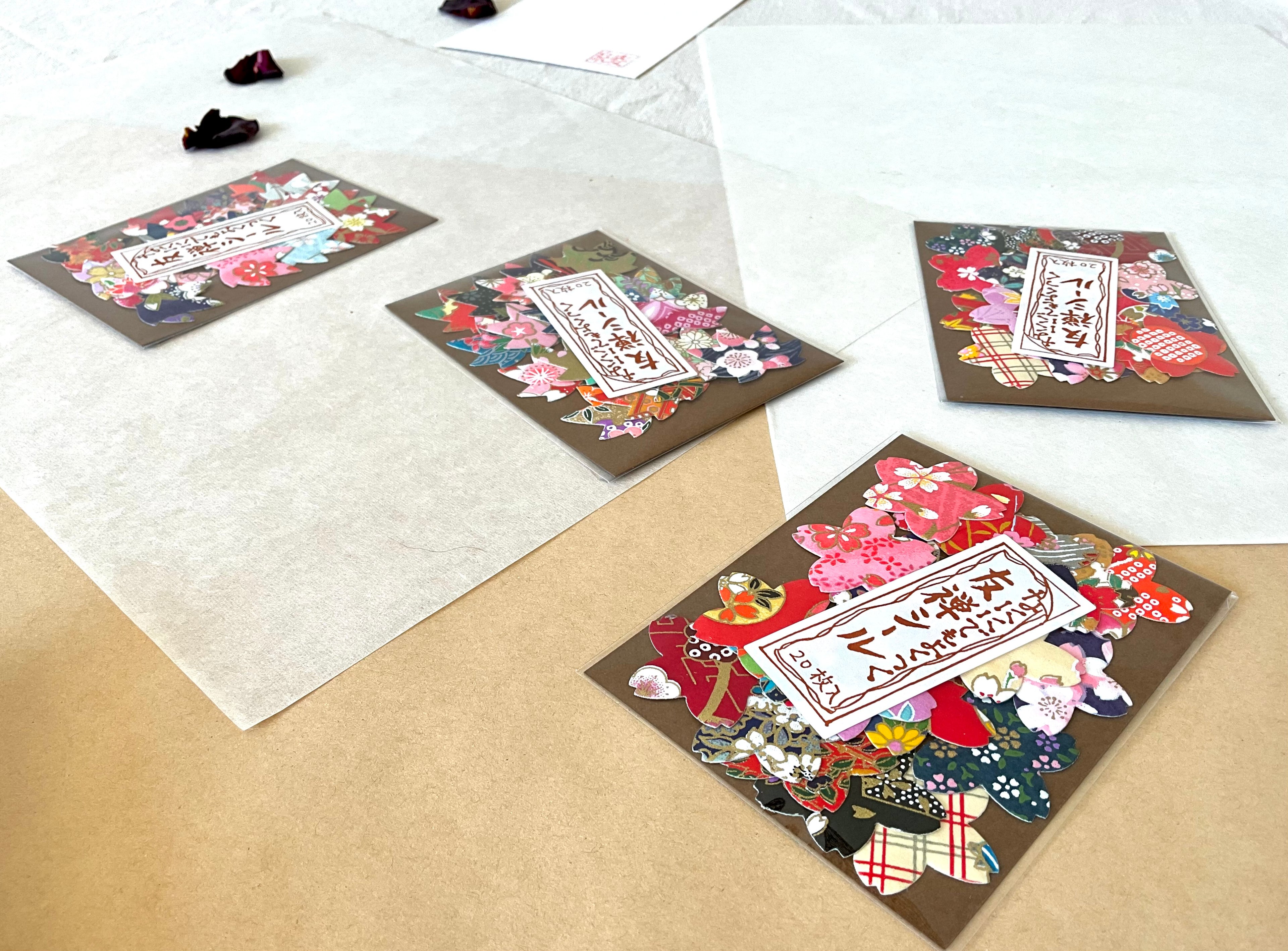Four packs of Morita Washi yuzen flower shaped stickers arranged at angles on layered washi and kraft paper, each pack showing a different assortment of colorful chiyogami petal cut stickers bursting out of brown card backing, with dried rose petals and hanko stamp card
