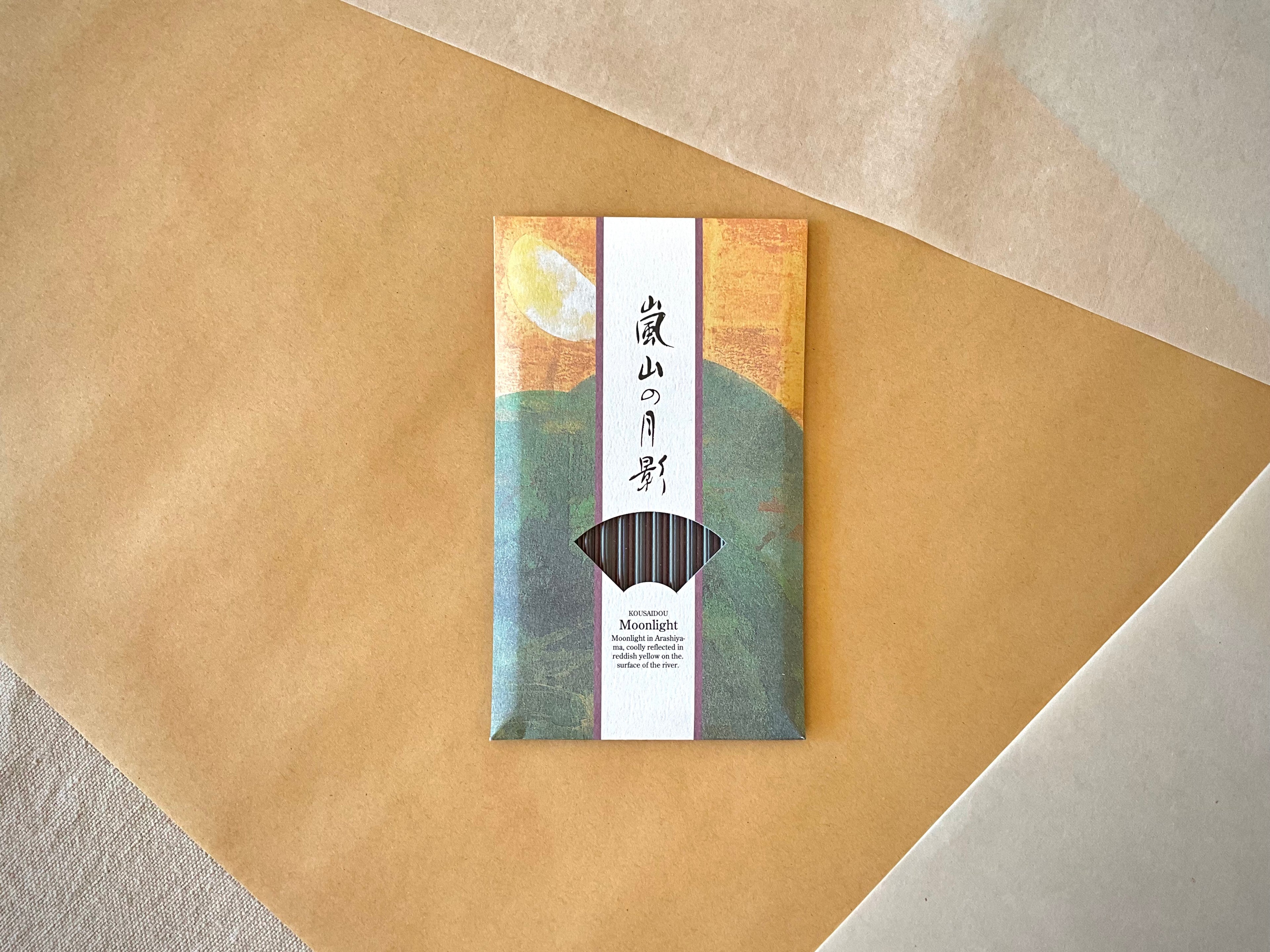 Kousaidou Arashiyama Moonlight incense pack centered on diagonal kraft paper background, full illustrated sleeve visible with Arashiyama mountain and moon design, fan window showing dark incense sticks, Kousaidou and Moonlight text in English