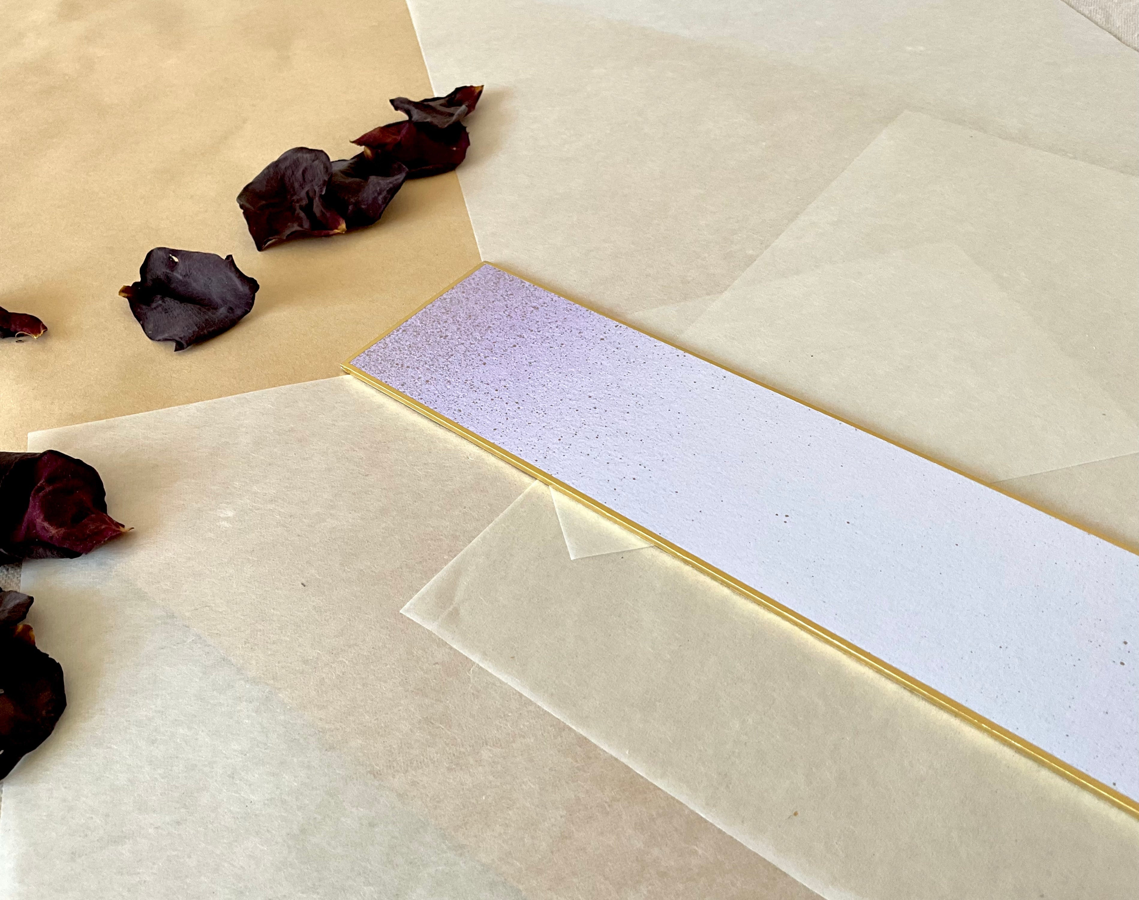 One Japanese tanzaku strips with gold edge, in lavender with fine speckled texture and , resting at an angle on layered washi and kraft paper with dried rose petals nearby