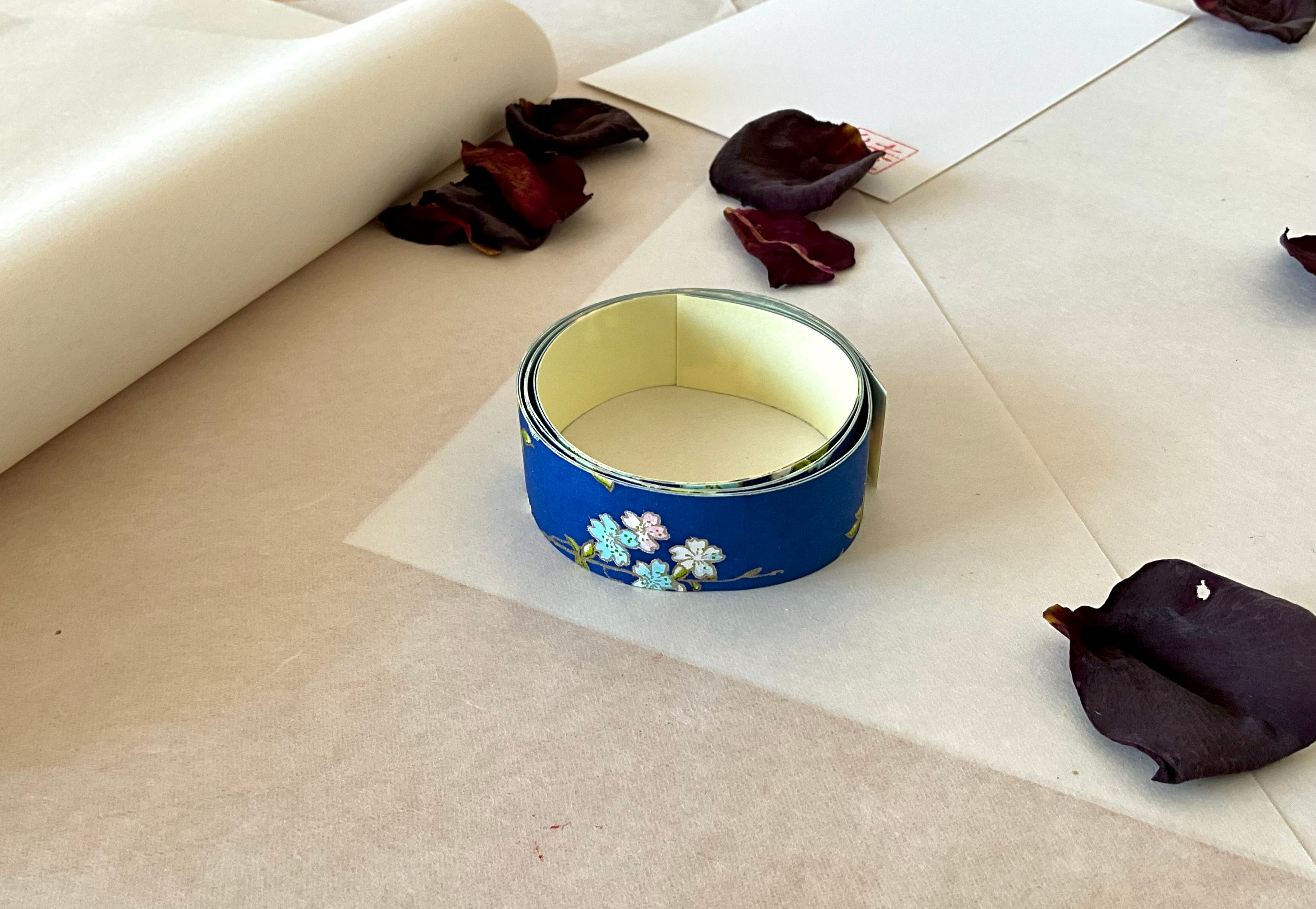 Shogado yuzen washi tape unrolled on washi paper showing Cobalt blue with white, pink and pale blue cherry blossom branches along its length, adhesive backing visible, dried rose petals nearby