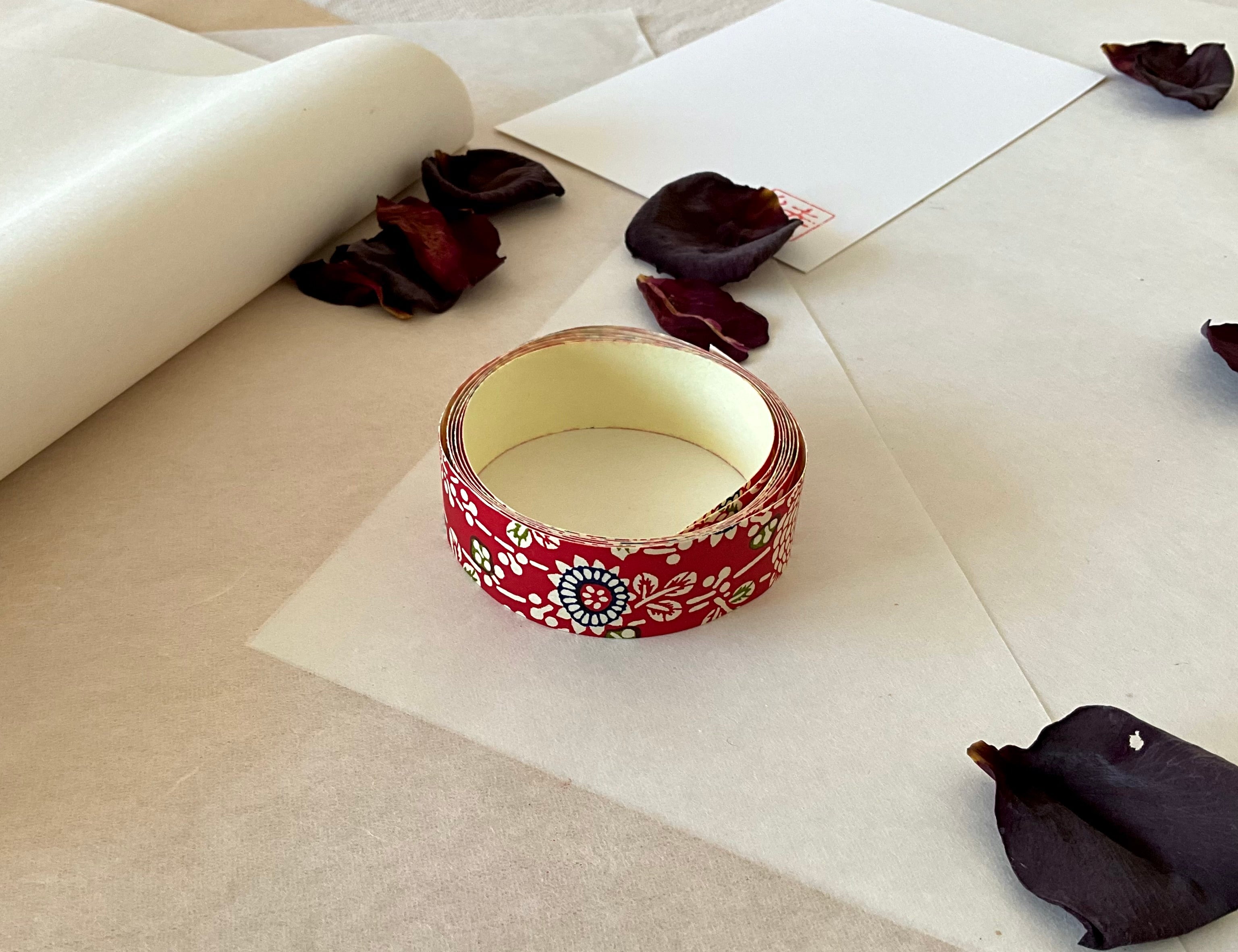 Shogado yuzen washi tape unrolled on washi paper showing Crimson red with cream chrysanthemum and floral motifs, navy dotted circle centers, green accents along its length, adhesive backing visible, dried rose petals nearby