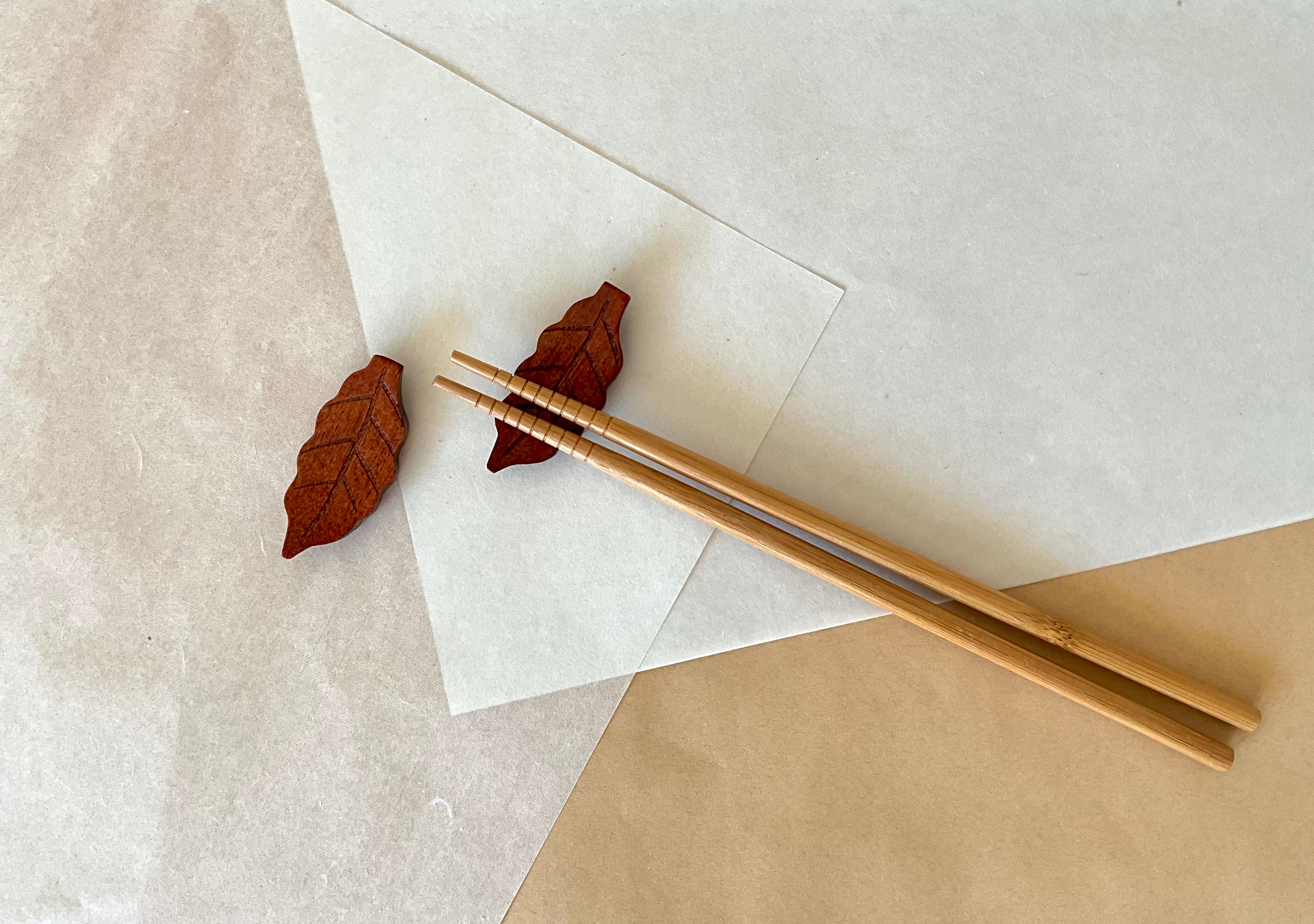 Pair of carved wood leaf chopstick rests holding bamboo chopsticks on neutral washi paper background