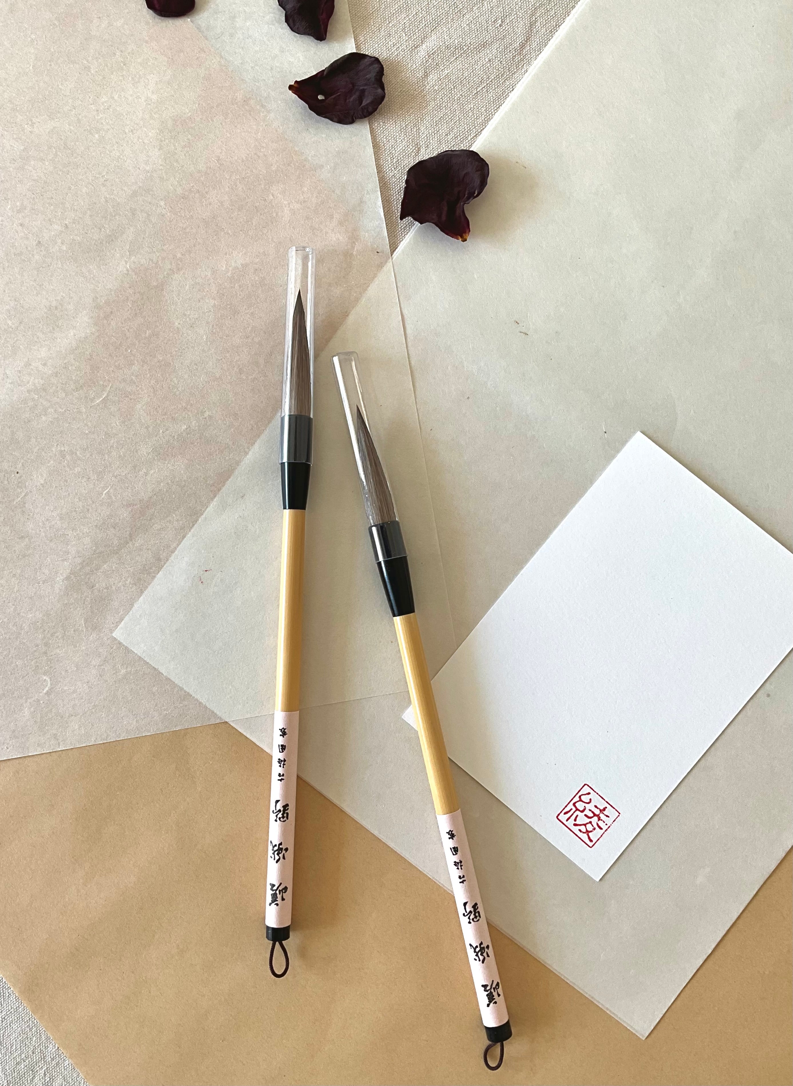 Two sealed Kobaien large calligraphy brushes with firm tapered bristle, black lacquered ferrule and pale bamboo handle with printed paper label and hanging loop, resting on layered washi paper with dried rose petals and a red hanko stamp card