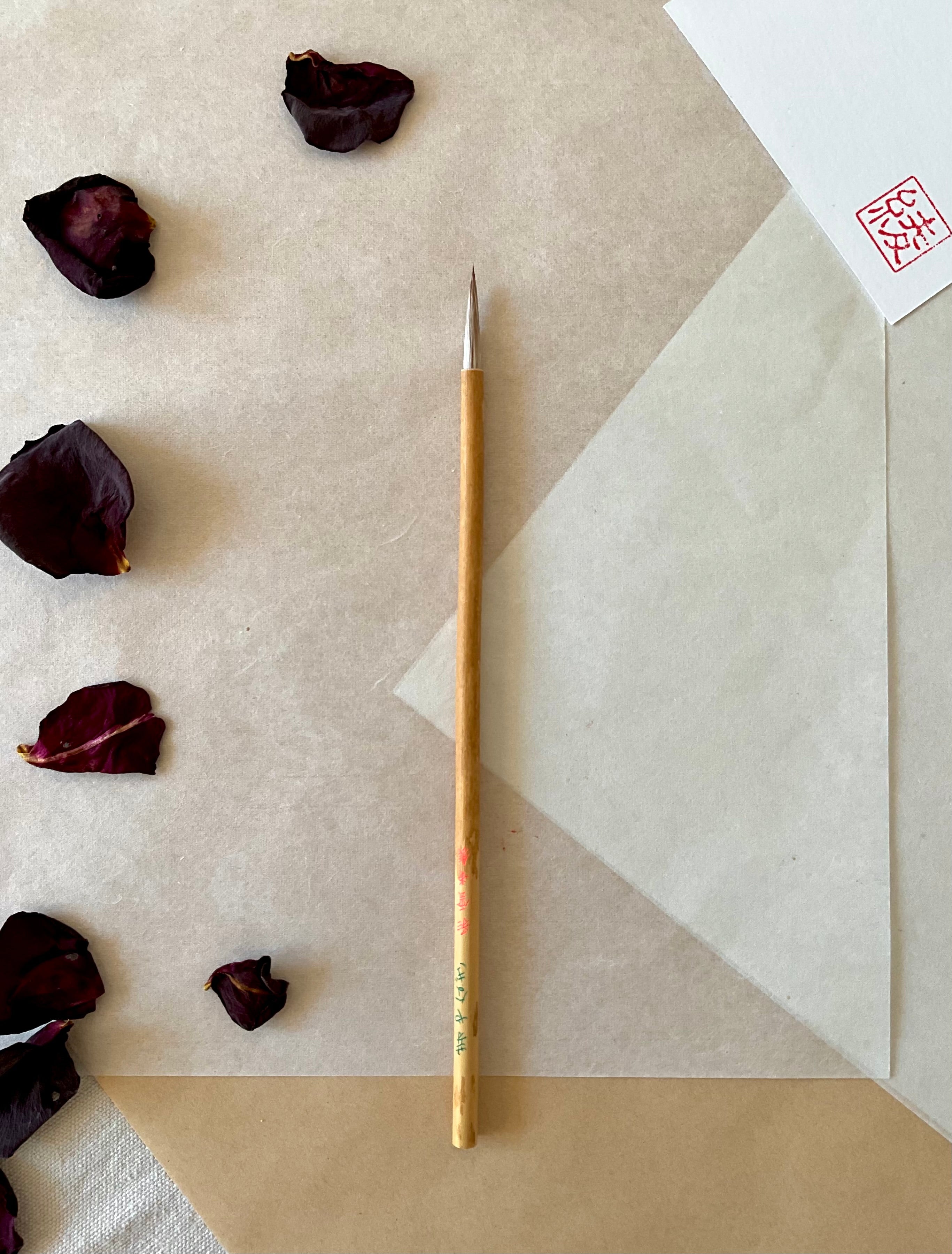 Saiundo calligraphy brush with light bamboo handle and fine pointed bristle, resting on layered washi paper with dried rose petals and a red hanko stamp card