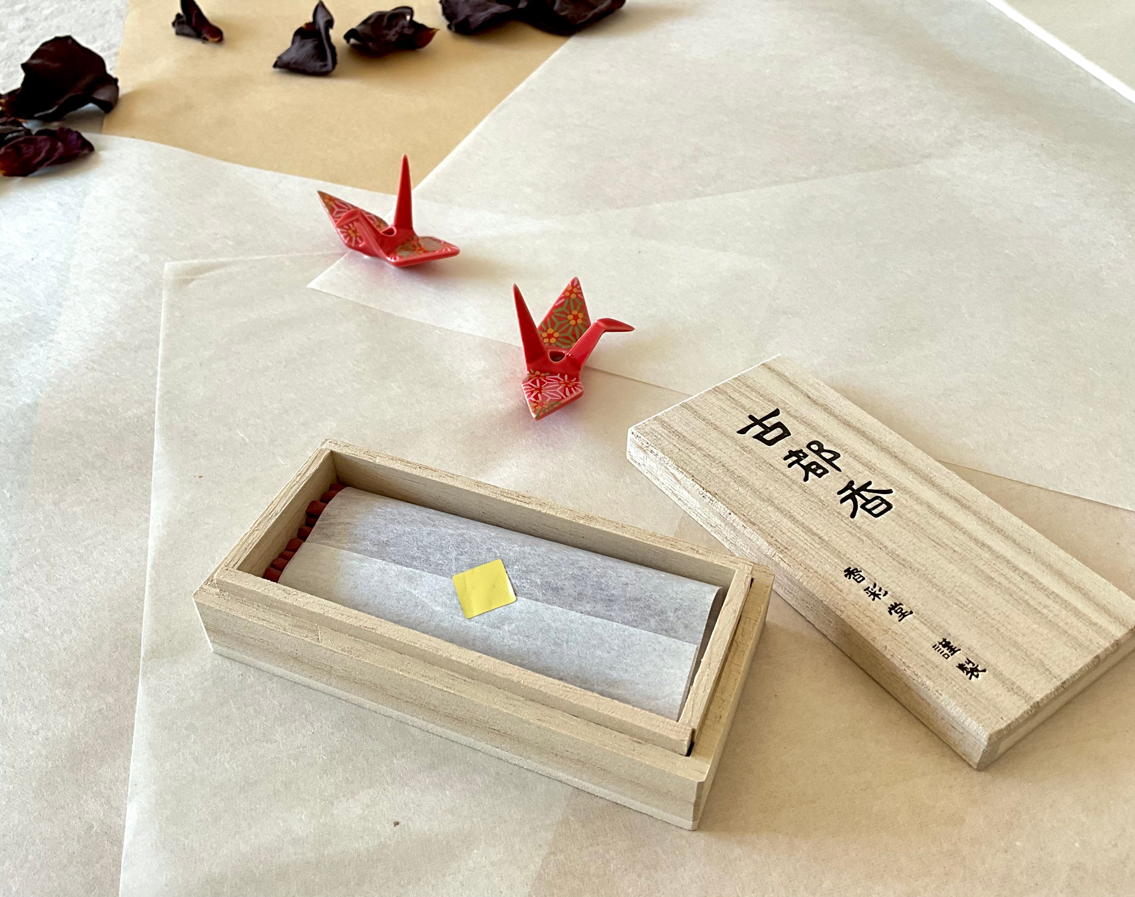 Kousaido Koto-ko incense paulownia wood box open showing incense sticks wrapped in tissue paper inside, lid beside it with Japanese kanji, two small red chiyogami origami cranes nearby on washi paper, dried rose petals in background