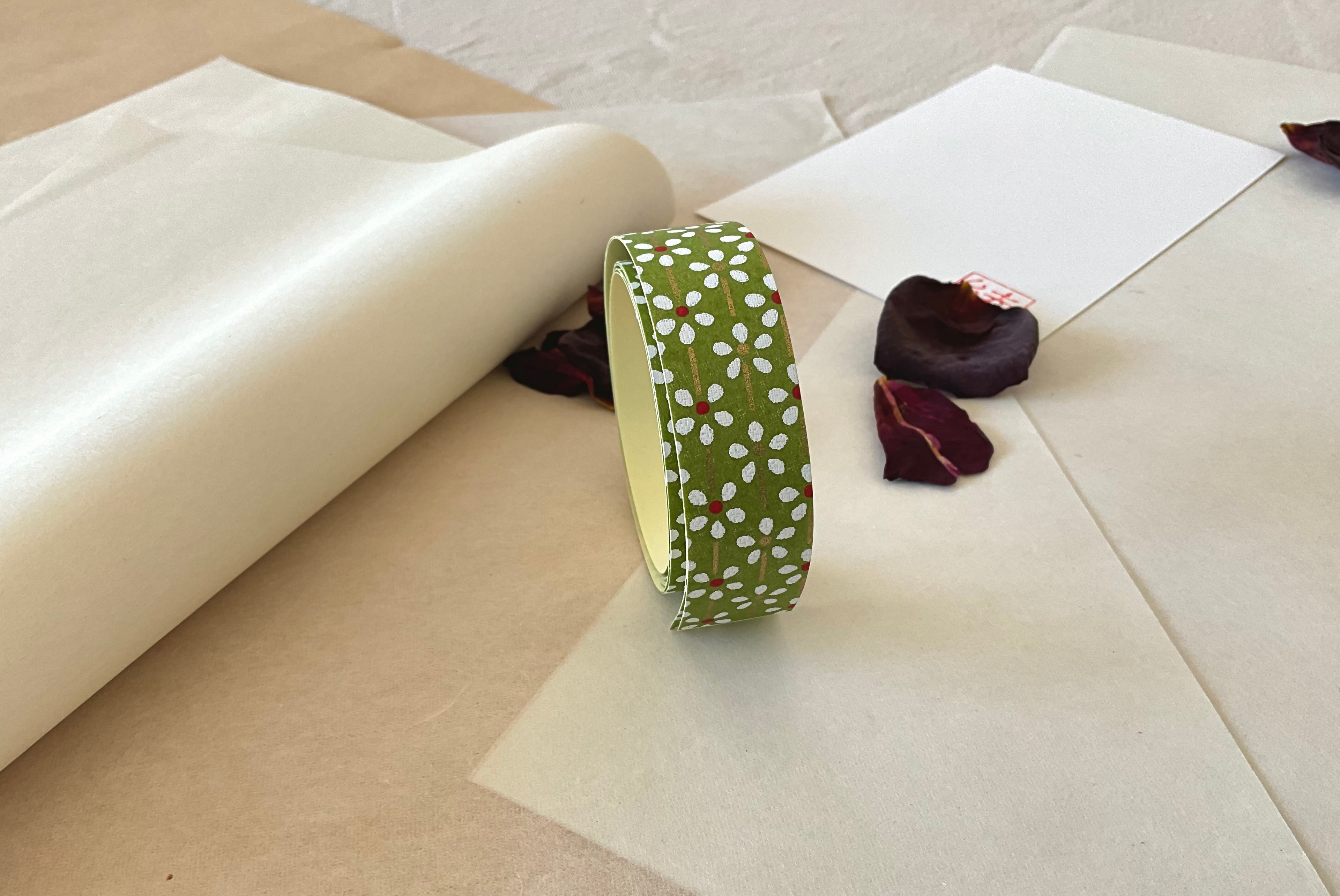 Roll of Shogado hand dyed yuzen washi tape standing upright on layered washi and kraft paper, Olive green with white daisy and red center floral print, cream adhesive backing visible, dried rose petals and hanko stamp card in background
