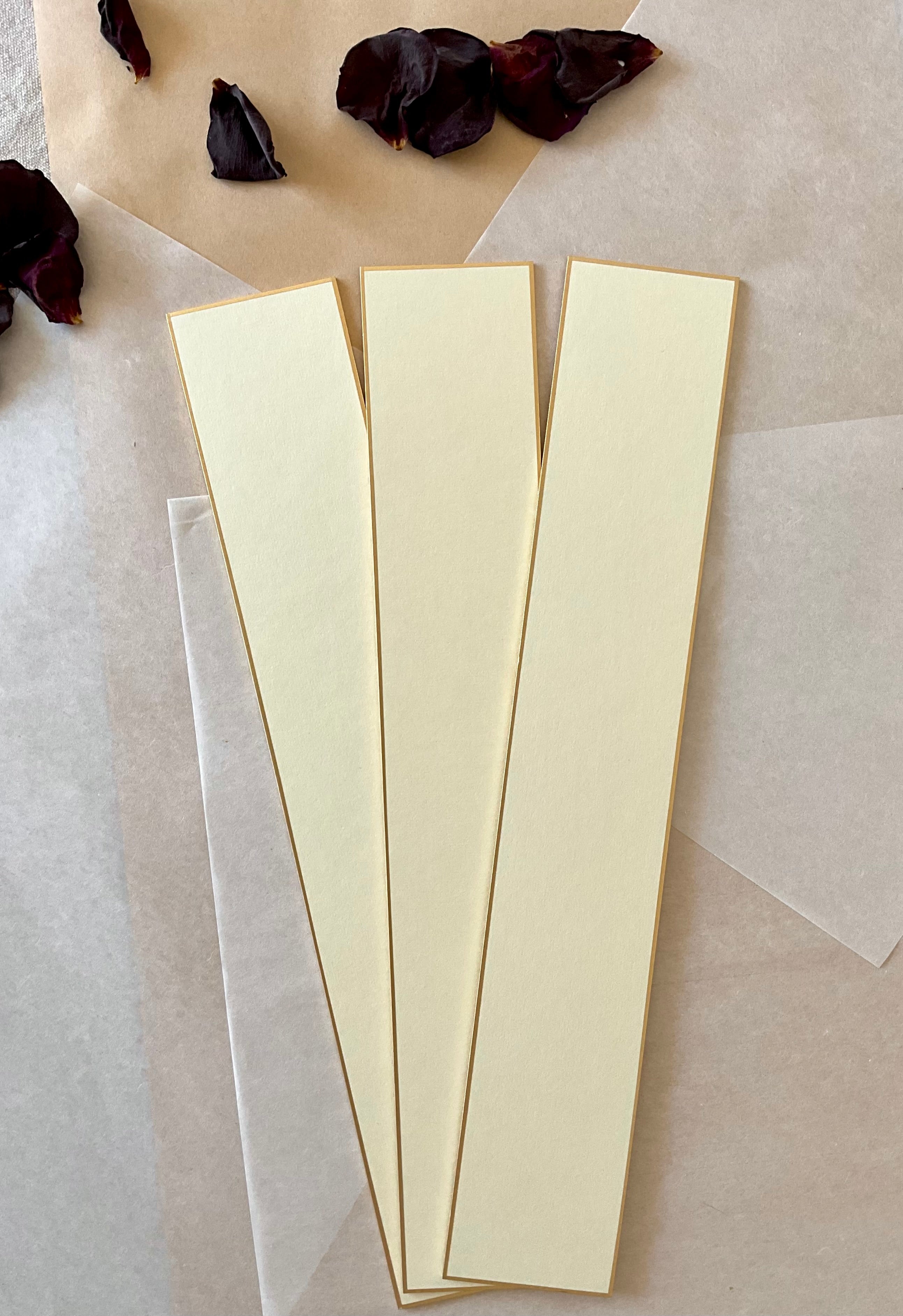 Three cream Kyukyodo tanzaku strips standing vertically side by side on washi paper with dried dark rose petals above, showing the full length and gold-painted edges