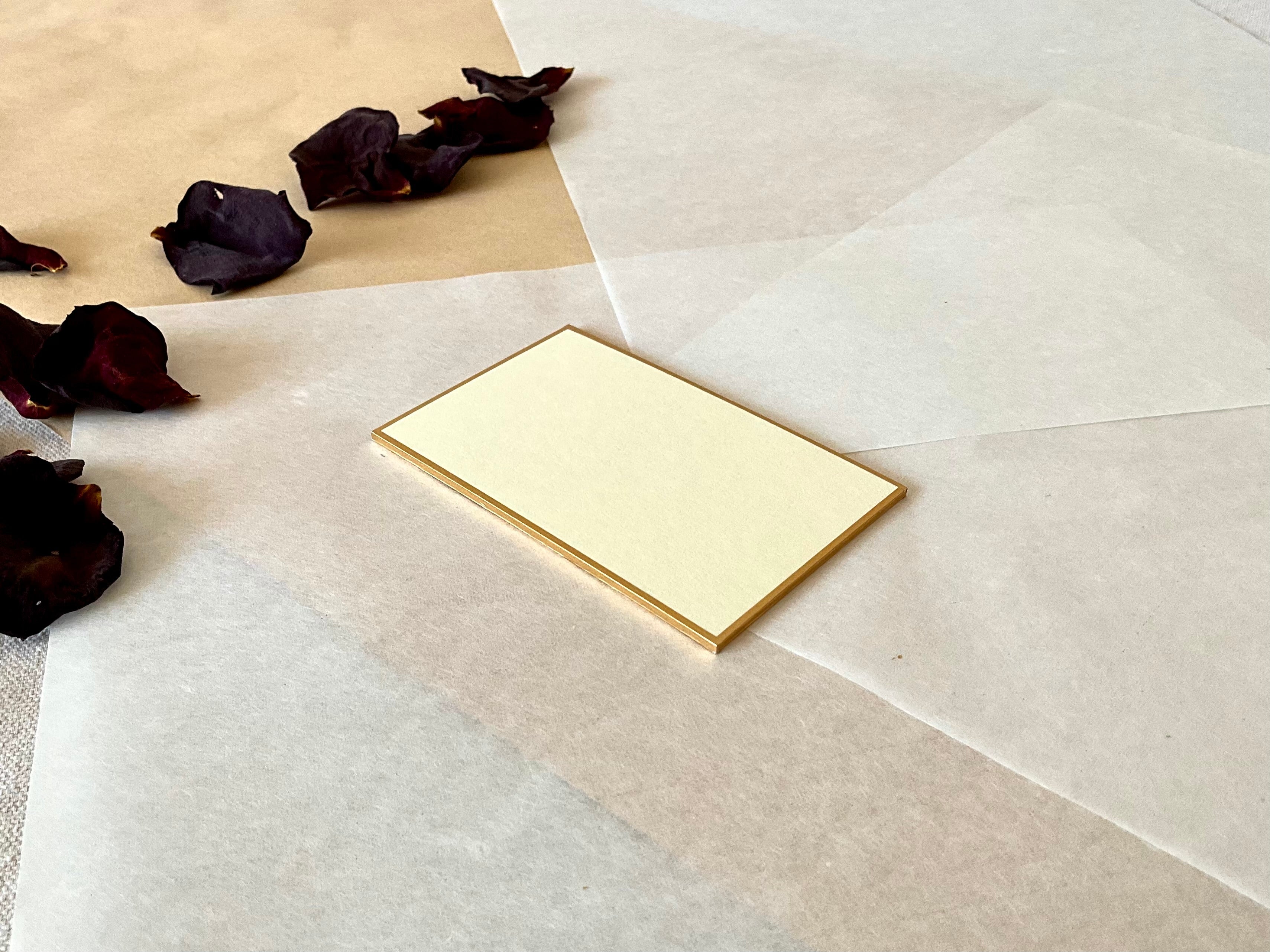 Single small rectangle cream Kyukyodo shikishi board from an angle layered on  washi and kraft paper with dried dark rose petals at upper left, showing the gold-painted edge