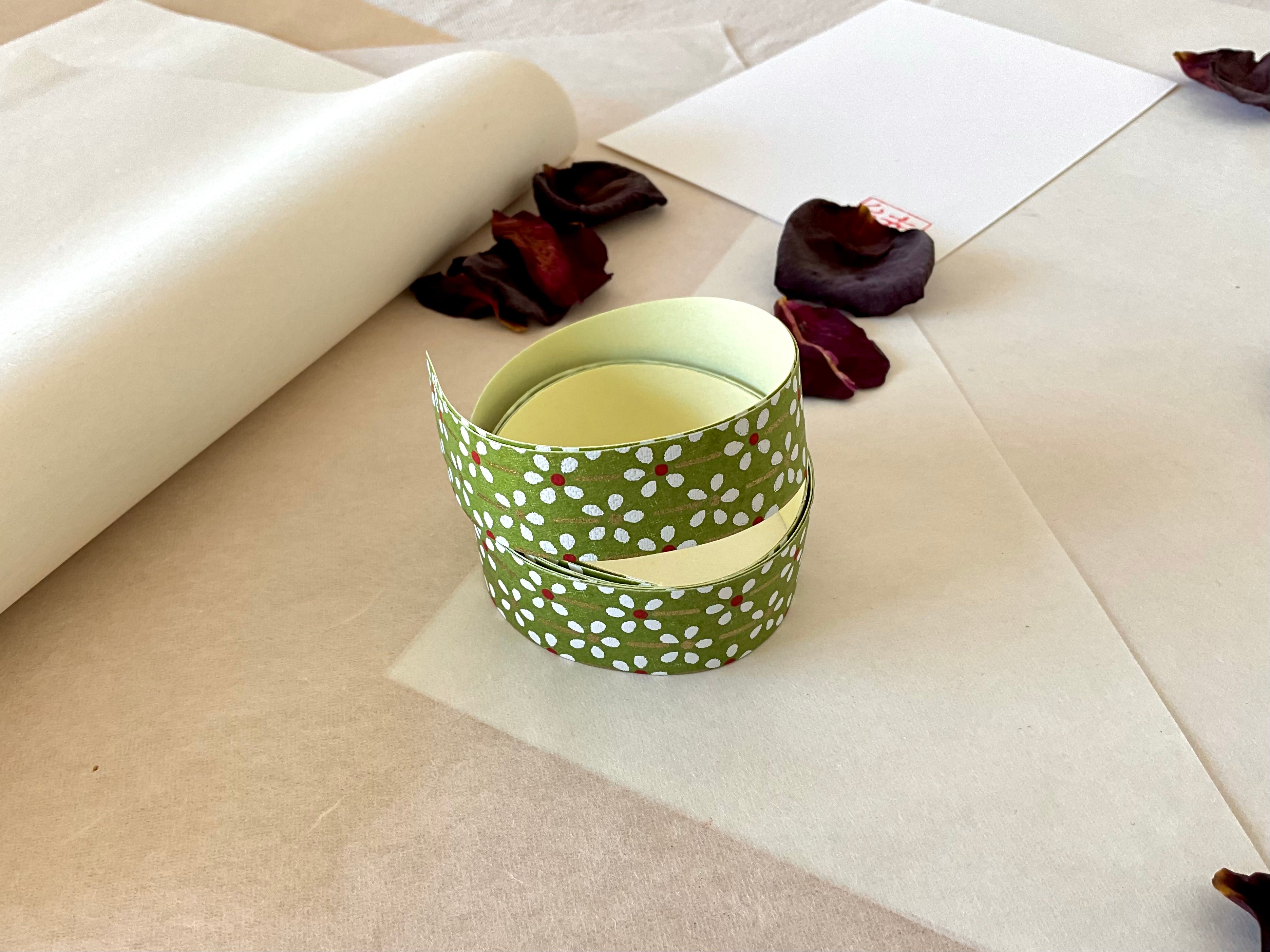 Both sheets of Shogado hand dyed yuzen washi tape laid on top of each other on  washi paper, showing Olive green with white daisy and red center floral print repeated across both strips, dried rose petals nearby