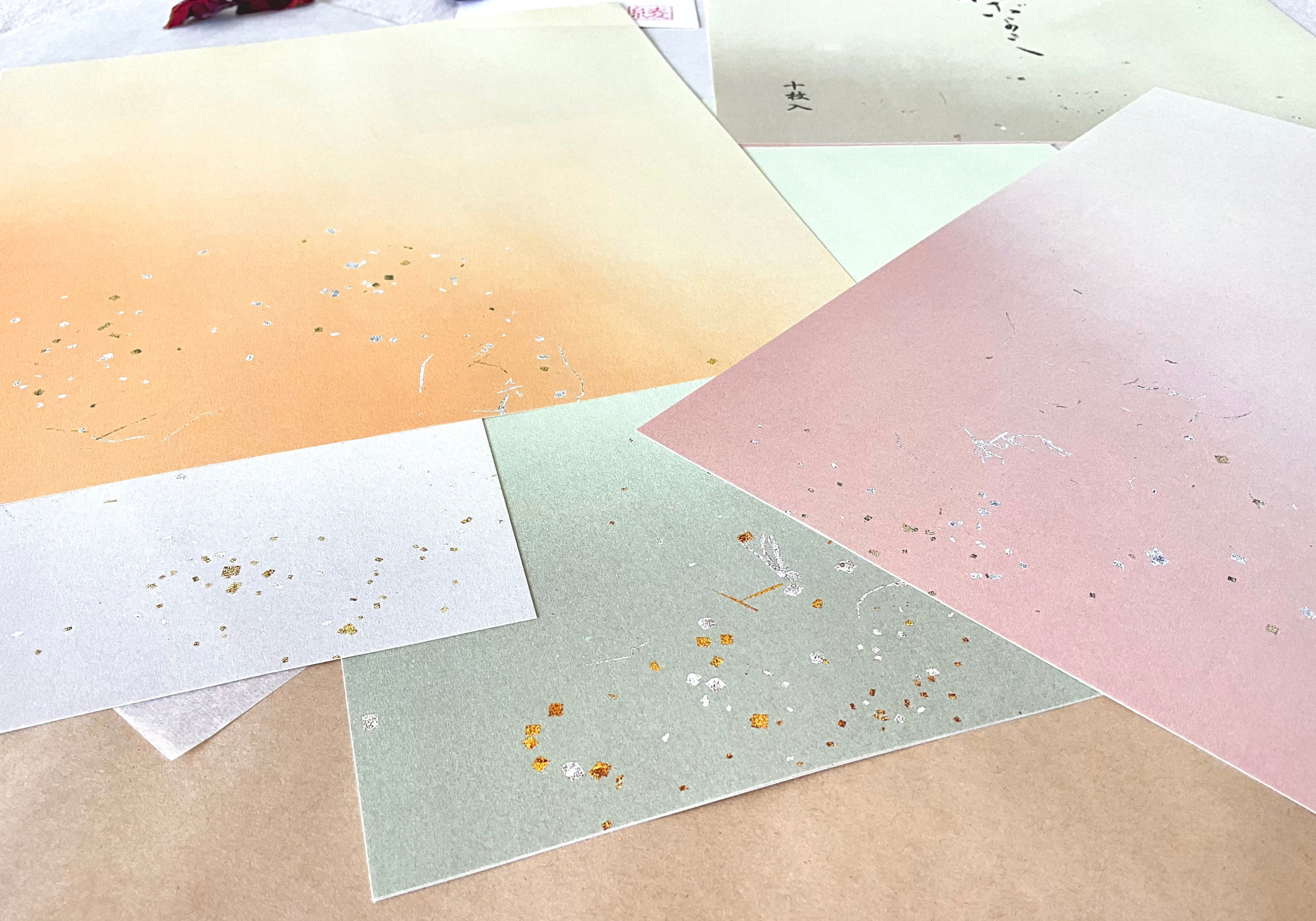 Close-up of four shikishi calligraphy sheets in orange, sage green, dusty rose and silver-grey, showing gold and silver fleck detail across the paper surface