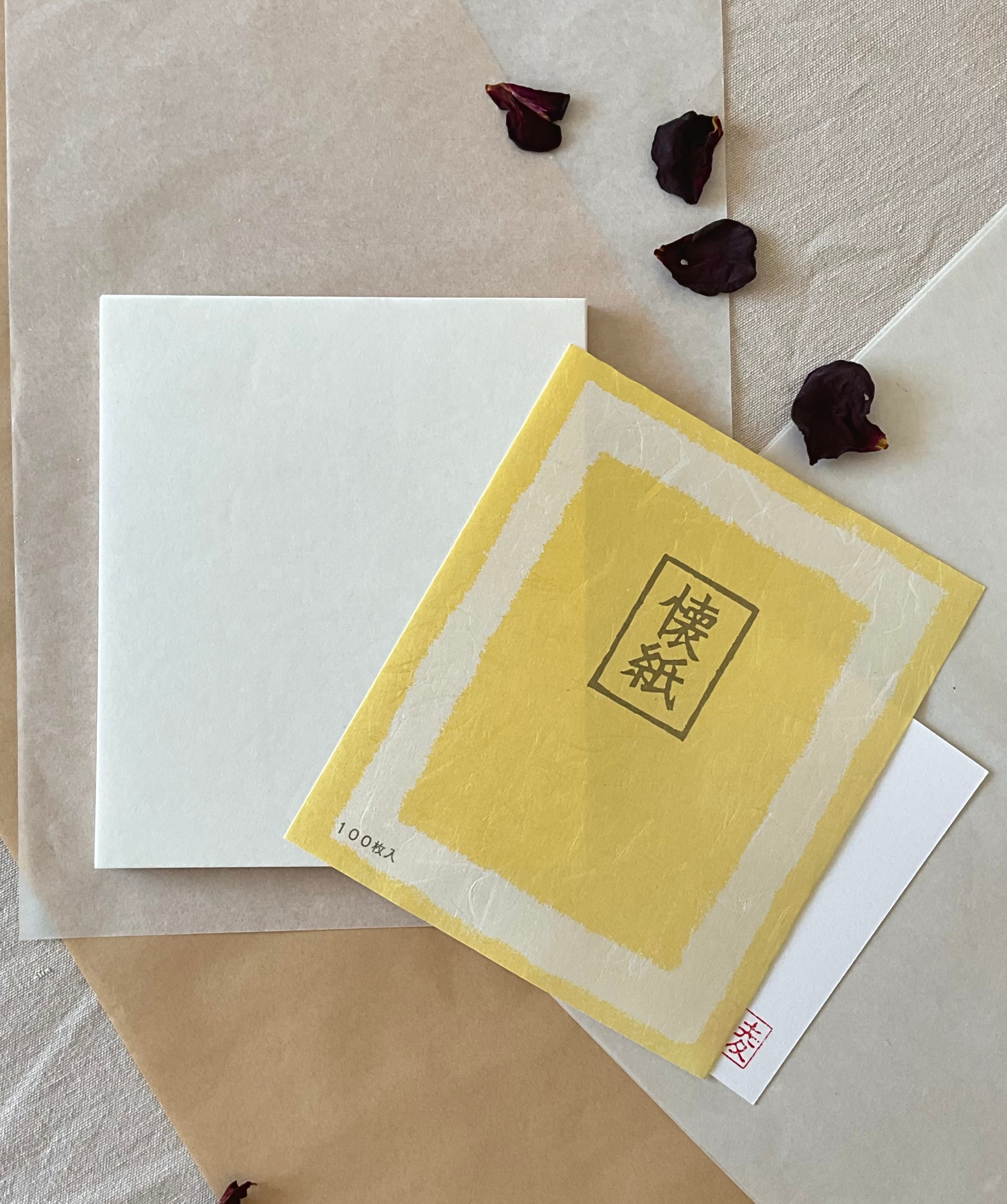 One packs of Morita Washi kaishi tea ceremony paper overlapping on layered washi and kraft paper, yellow washi covers with Japanese kaishi characters, 100 sheets each, with dried rose petals and hanko stamp card