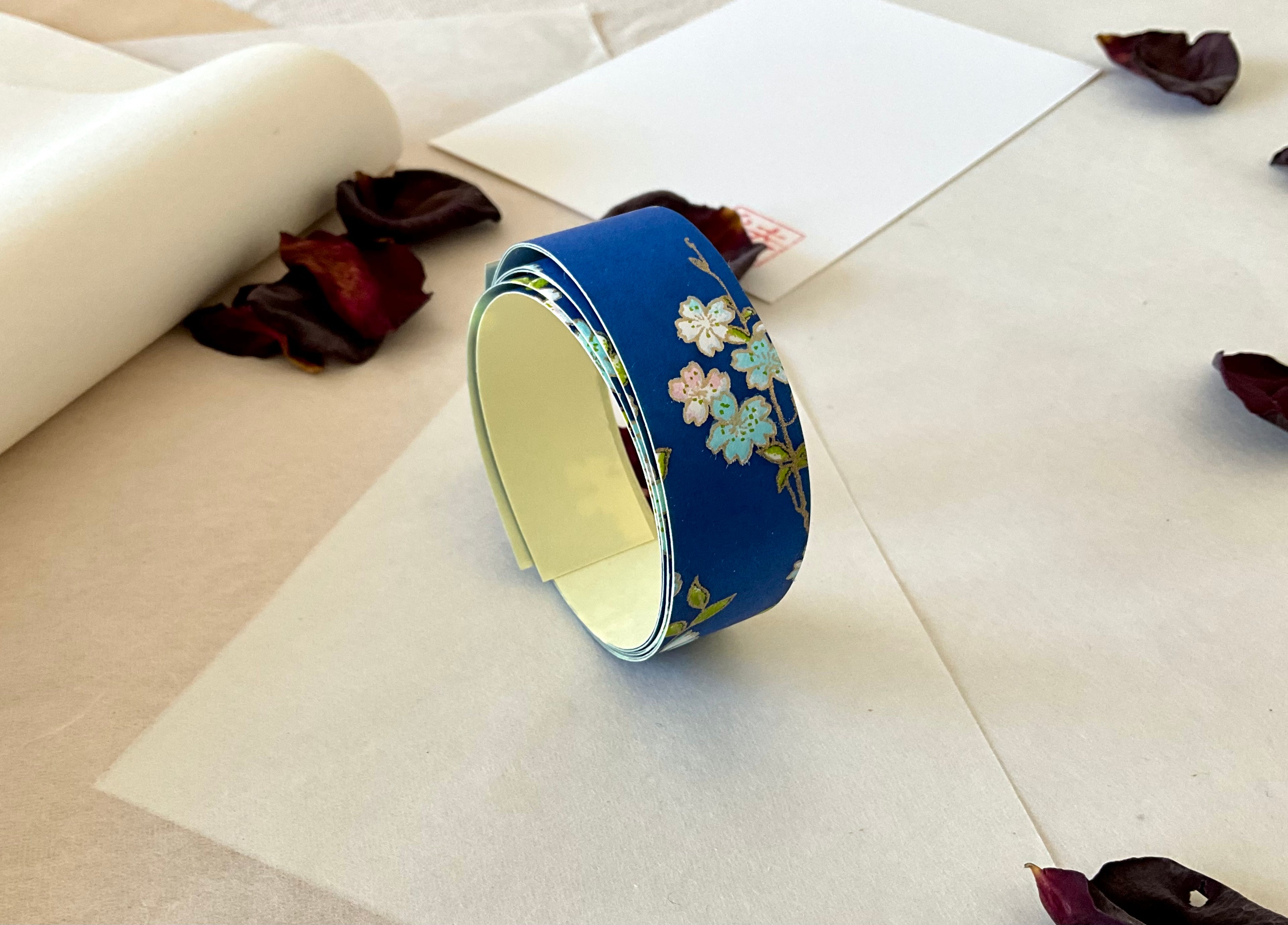 Roll of Shogado hand dyed yuzen washi tape standing upright on layered washi and kraft paper, Cobalt blue with white, pink and pale blue cherry blossom branches, cream adhesive backing visible, dried rose petals and hanko stamp card in background