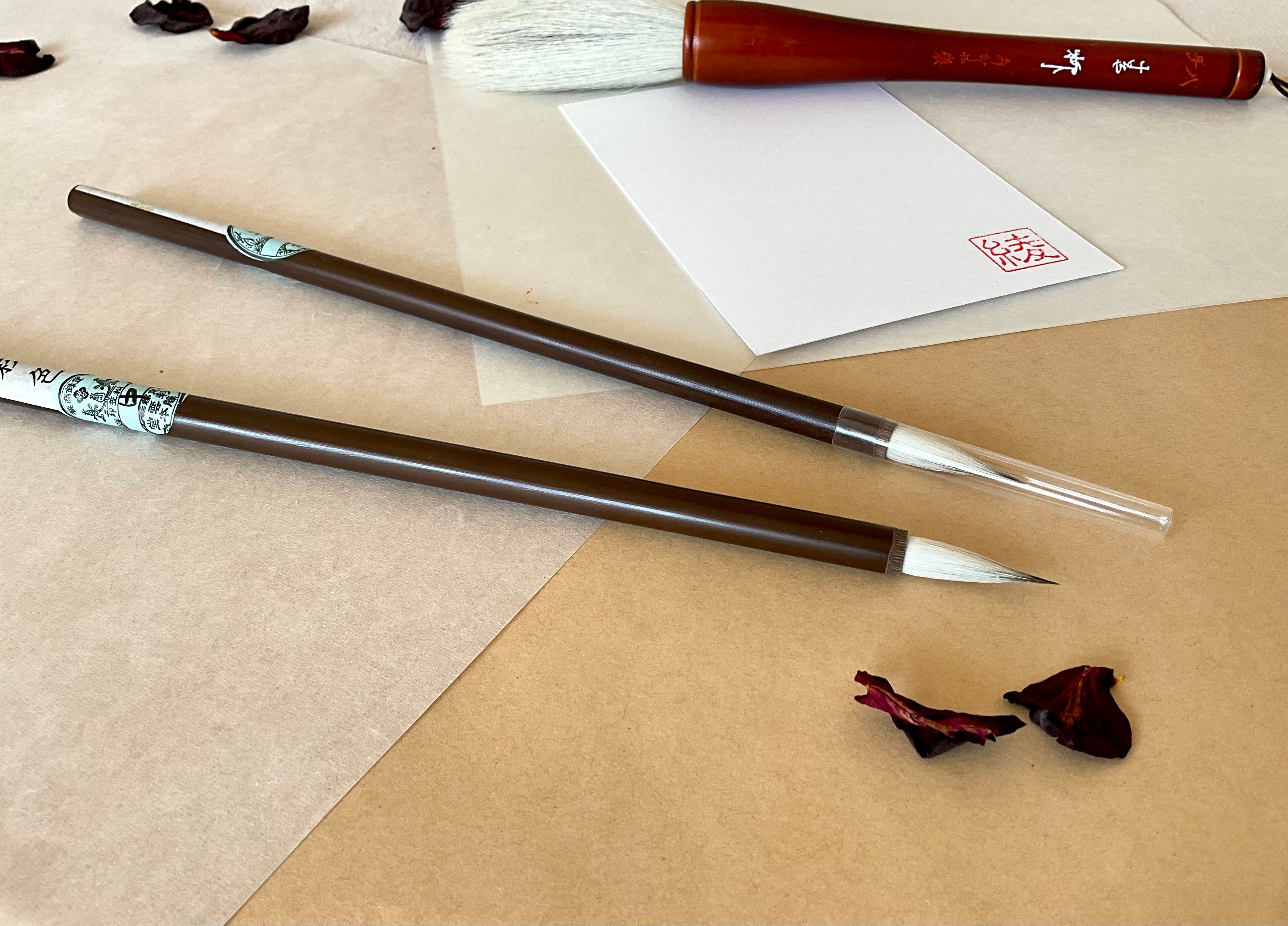 Two Saiundo calligraphy brush with dark lacquered handle and printed paper label, full pointed bristle, one is covered with a lid and one is open, resting on layered washi paper with dried rose petals and a red hanko stamp card