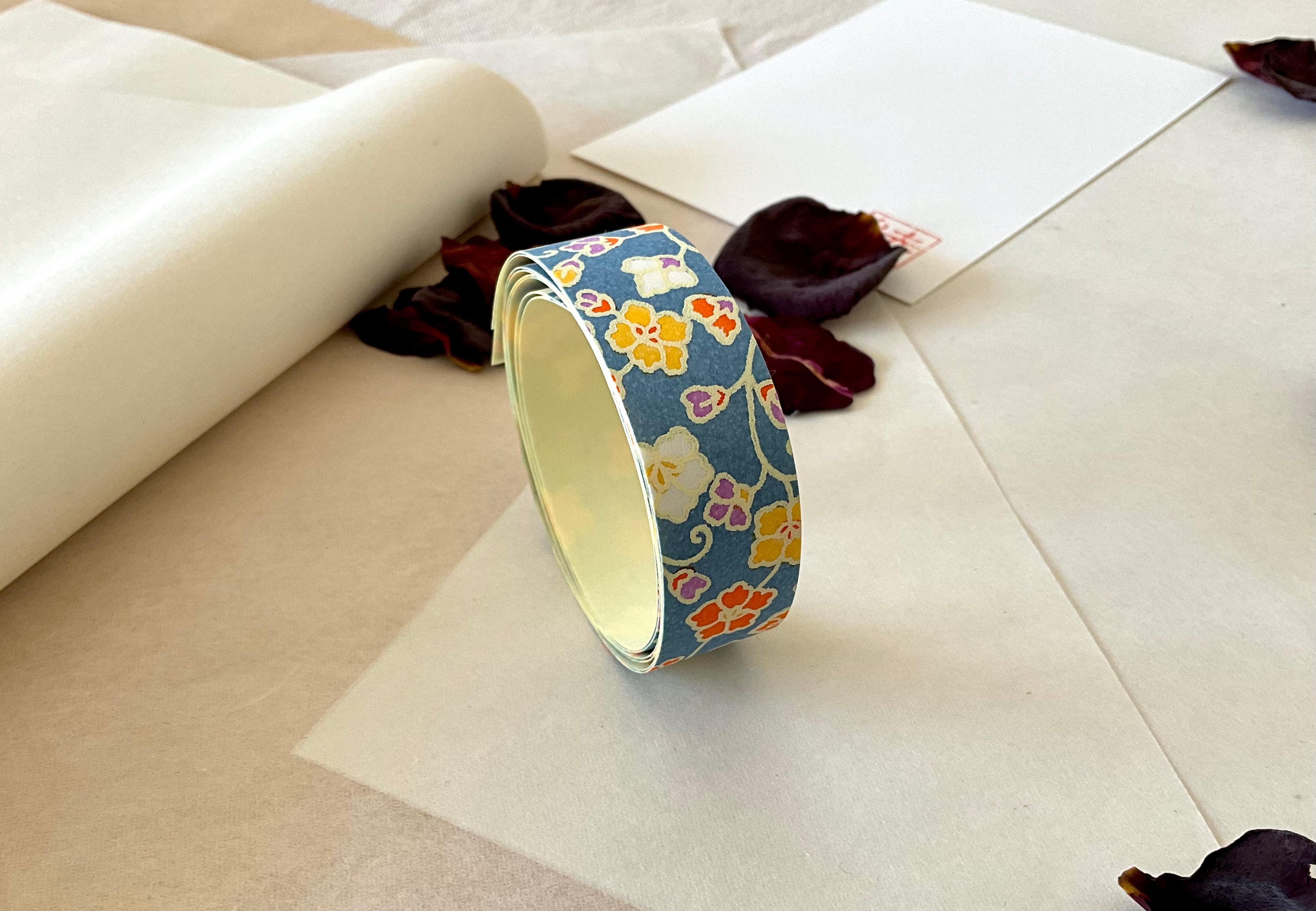 Roll of Shogado hand dyed yuzen washi tape standing upright on layered washi and kraft paper, Steel blue with yellow, orange, purple small florals, cream adhesive backing visible, dried rose petals and hanko stamp card in background