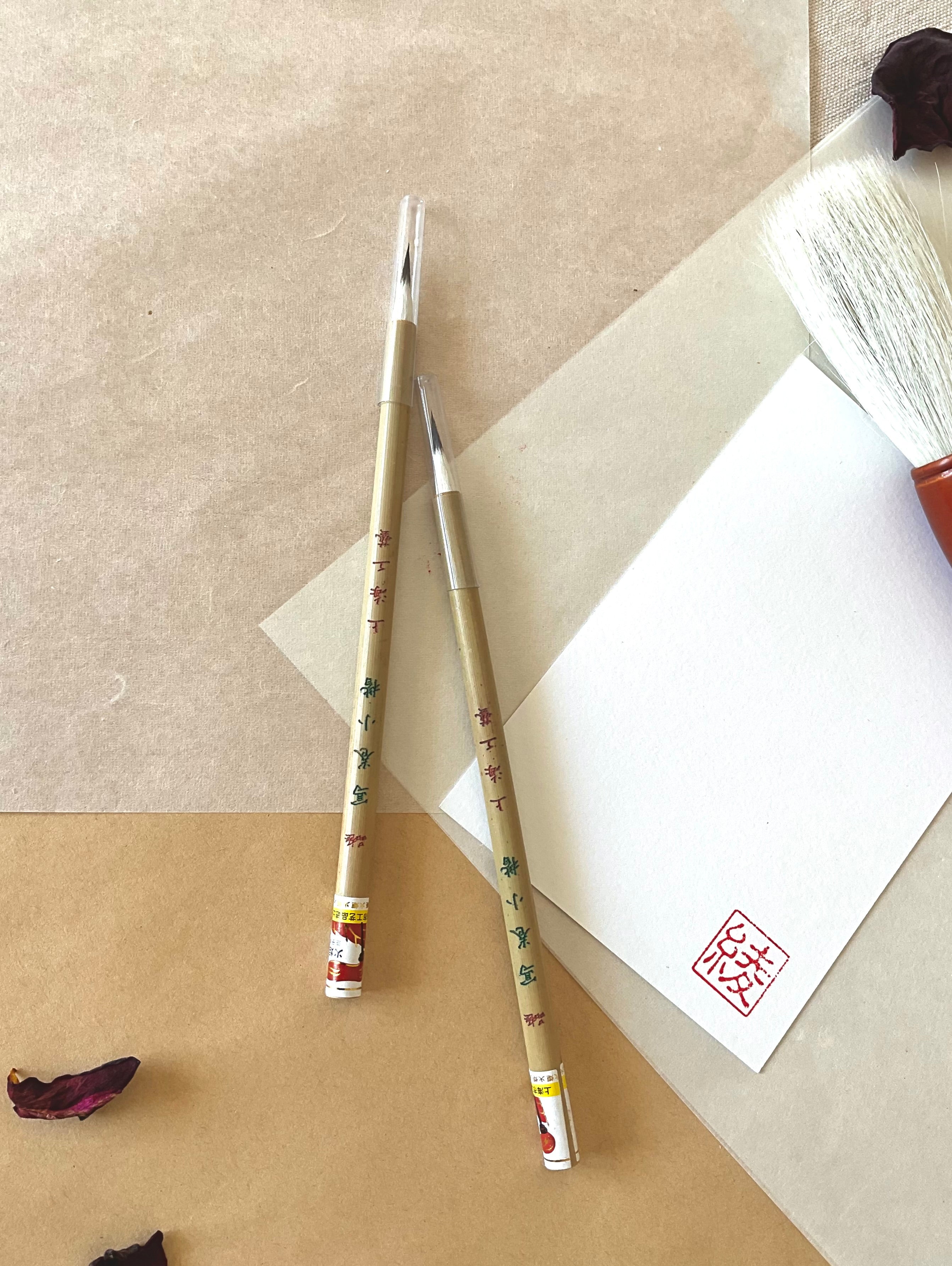 Two Kobaien fine detail calligraphy brushes with sharp pointed tip and bamboo handle printed with Japanese maker markings, resting on layered washi paper with dried rose petals and a red hanko stamp card