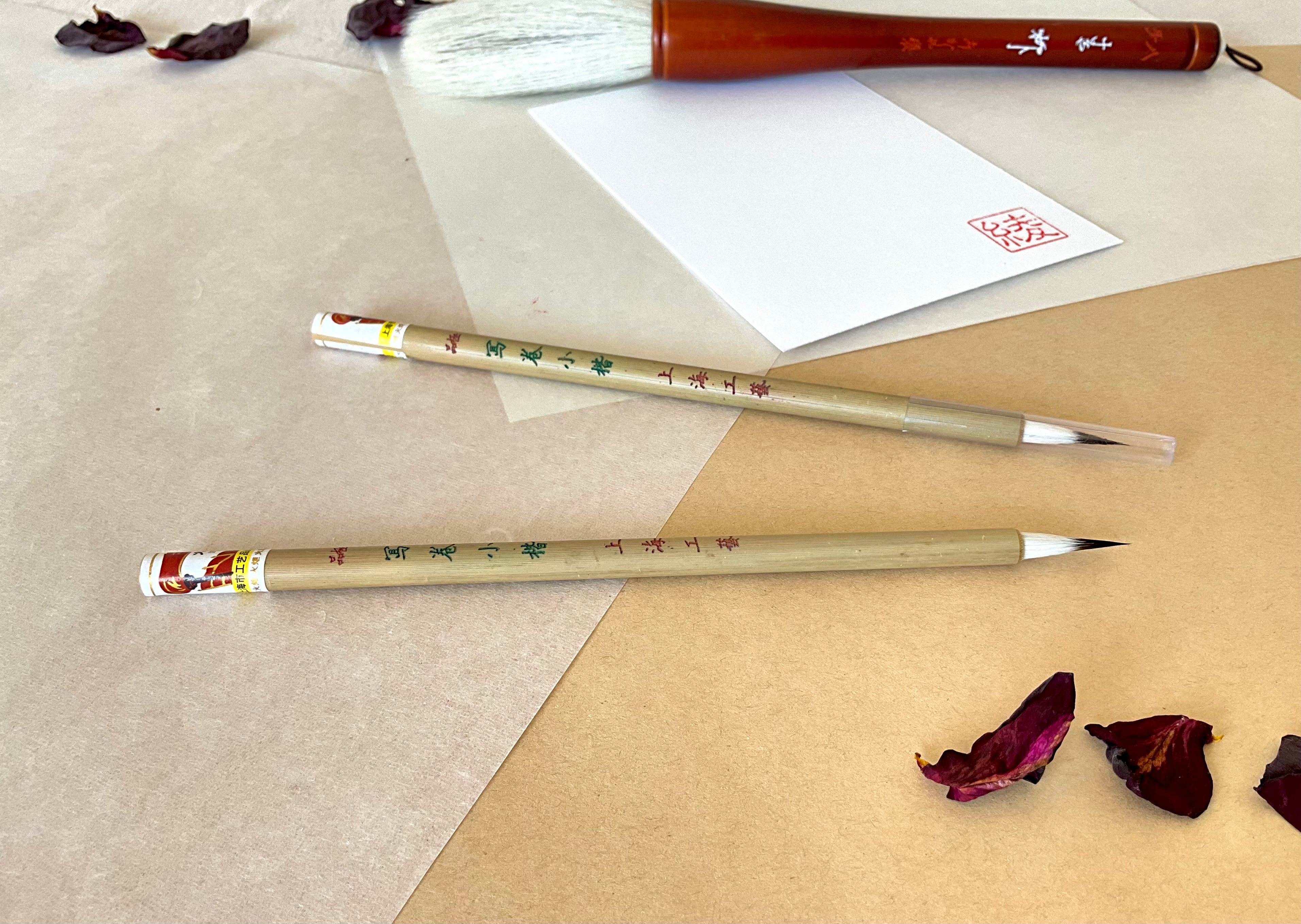 Two Kobaien fine detail calligraphy brushes with sharp pointed tip and bamboo handle printed with Japanese maker markings, one is covered with a lid and one is open, resting on layered washi paper with dried rose petals and a red hanko stamp card