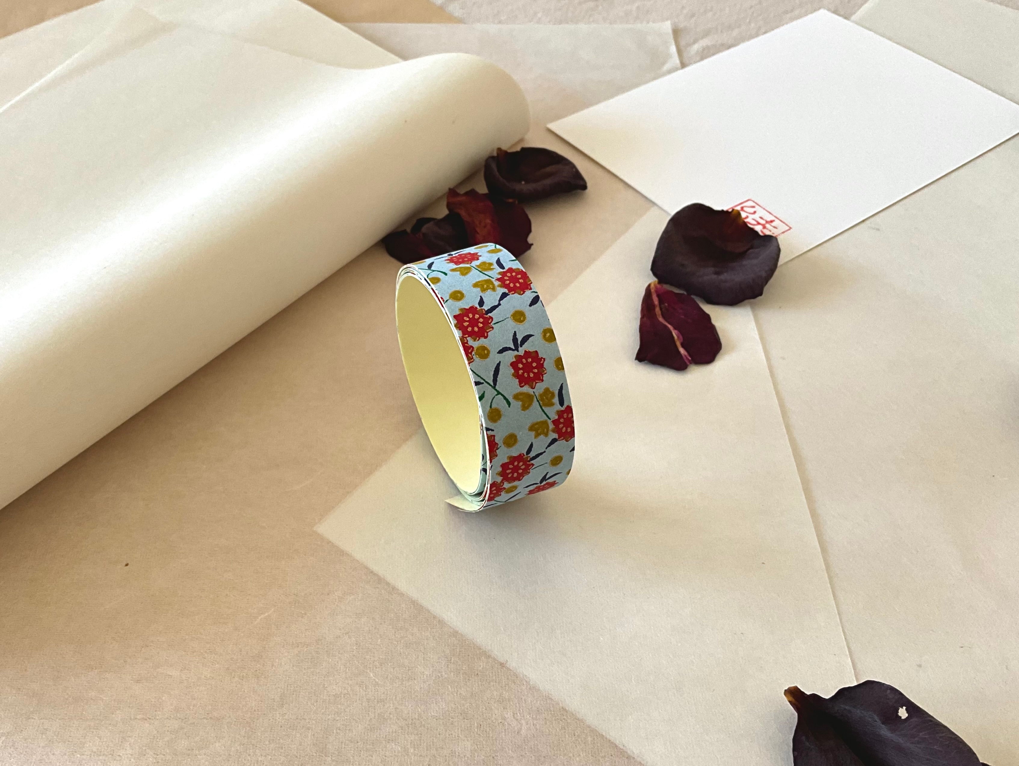 Roll of Shogado hand dyed yuzen washi tape standing upright on layered washi and kraft paper, Pale blue with red flowers, mustard dots, navy leaves, cream adhesive backing visible, dried rose petals and hanko stamp card in background