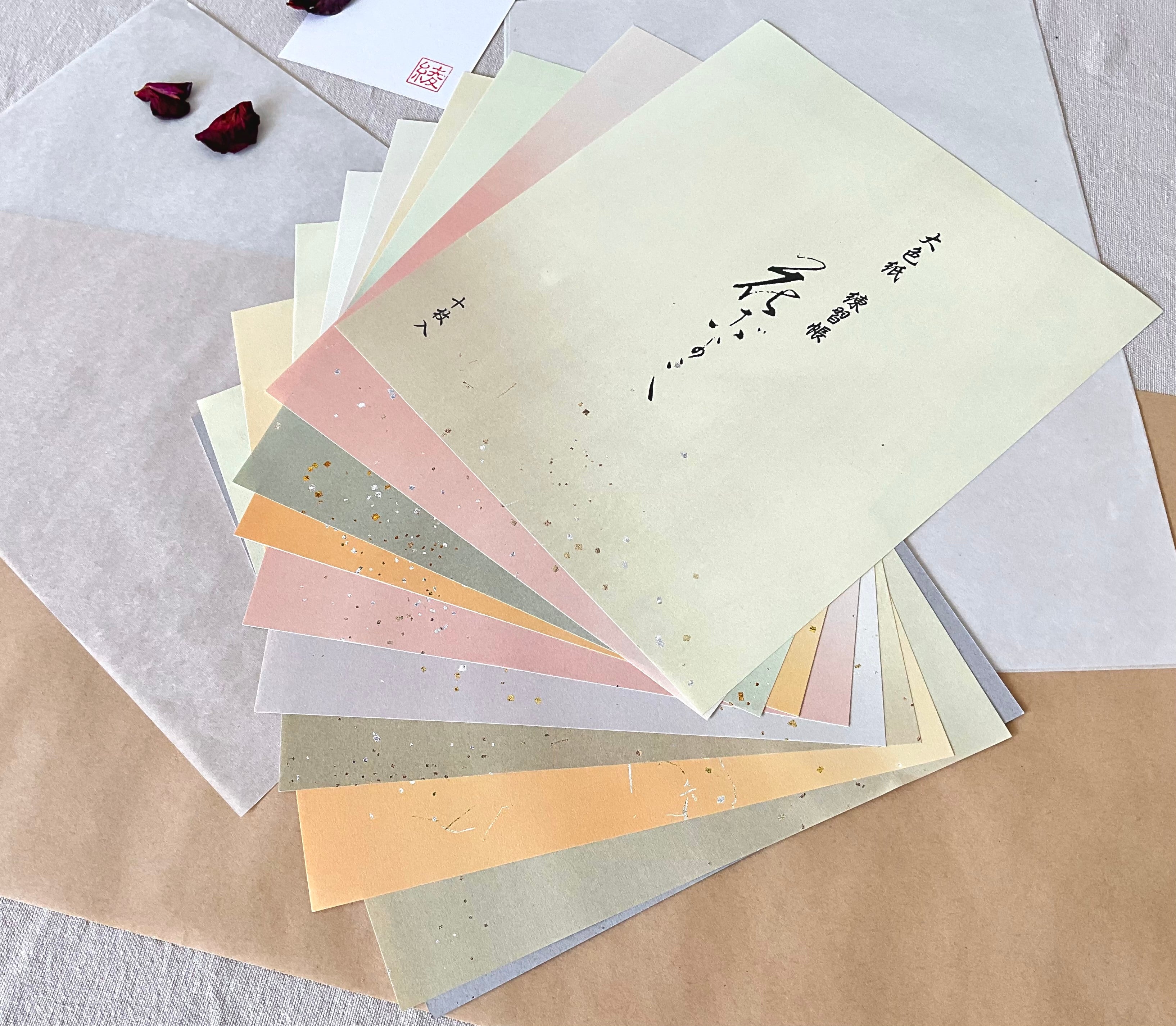 Ten colored shikishi calligraphy sheets fanned out showing full color range, sage, blush, cream, gold, grey- each with gold and silver metallic flecks, sourced from Kyoto Japan