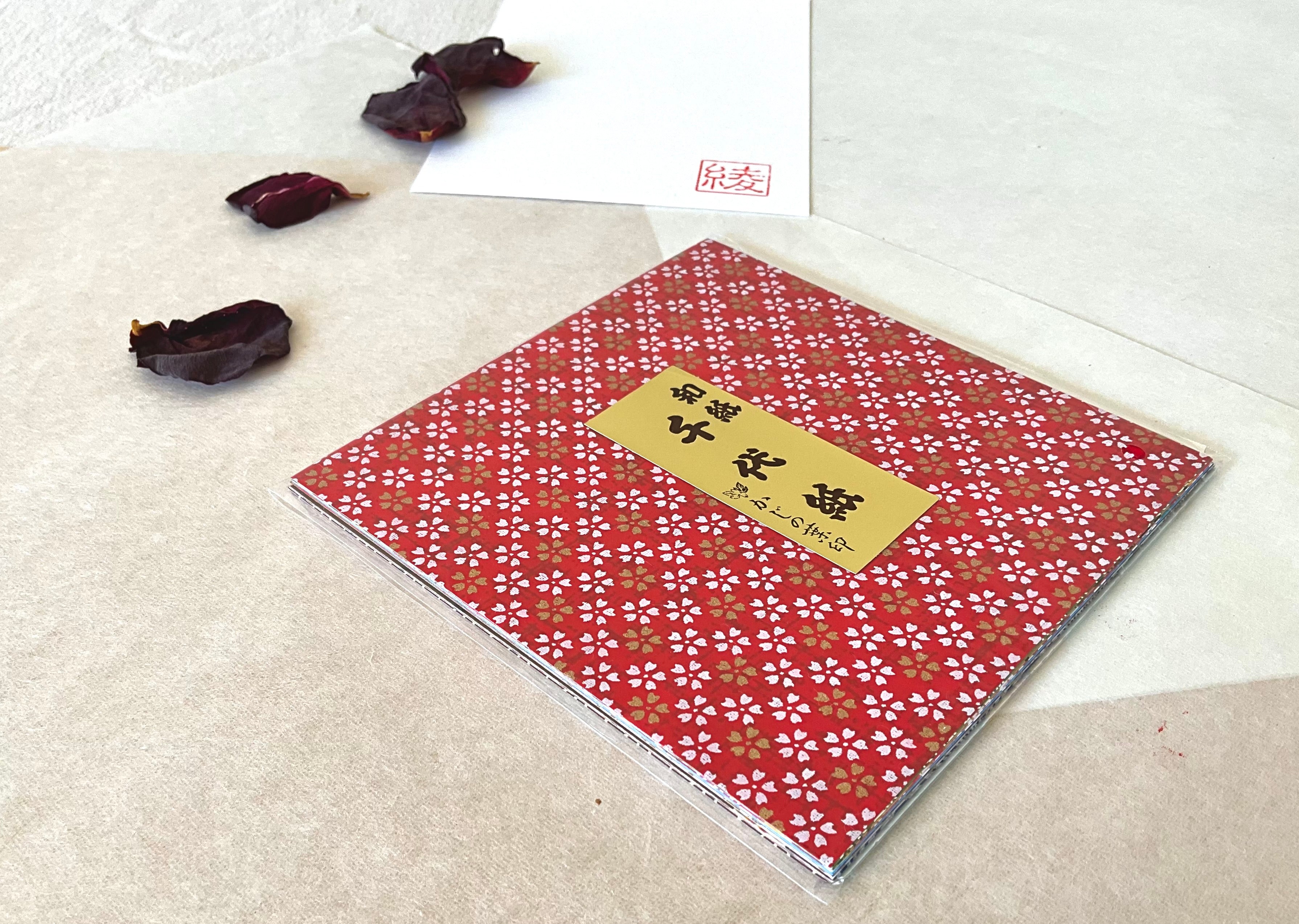 Morita Washi chiyogami origami paper pack at an angle showing depth of 20 sheets inside clear sleeve, deep red sakura cover pattern, on washi paper background with dried rose petals