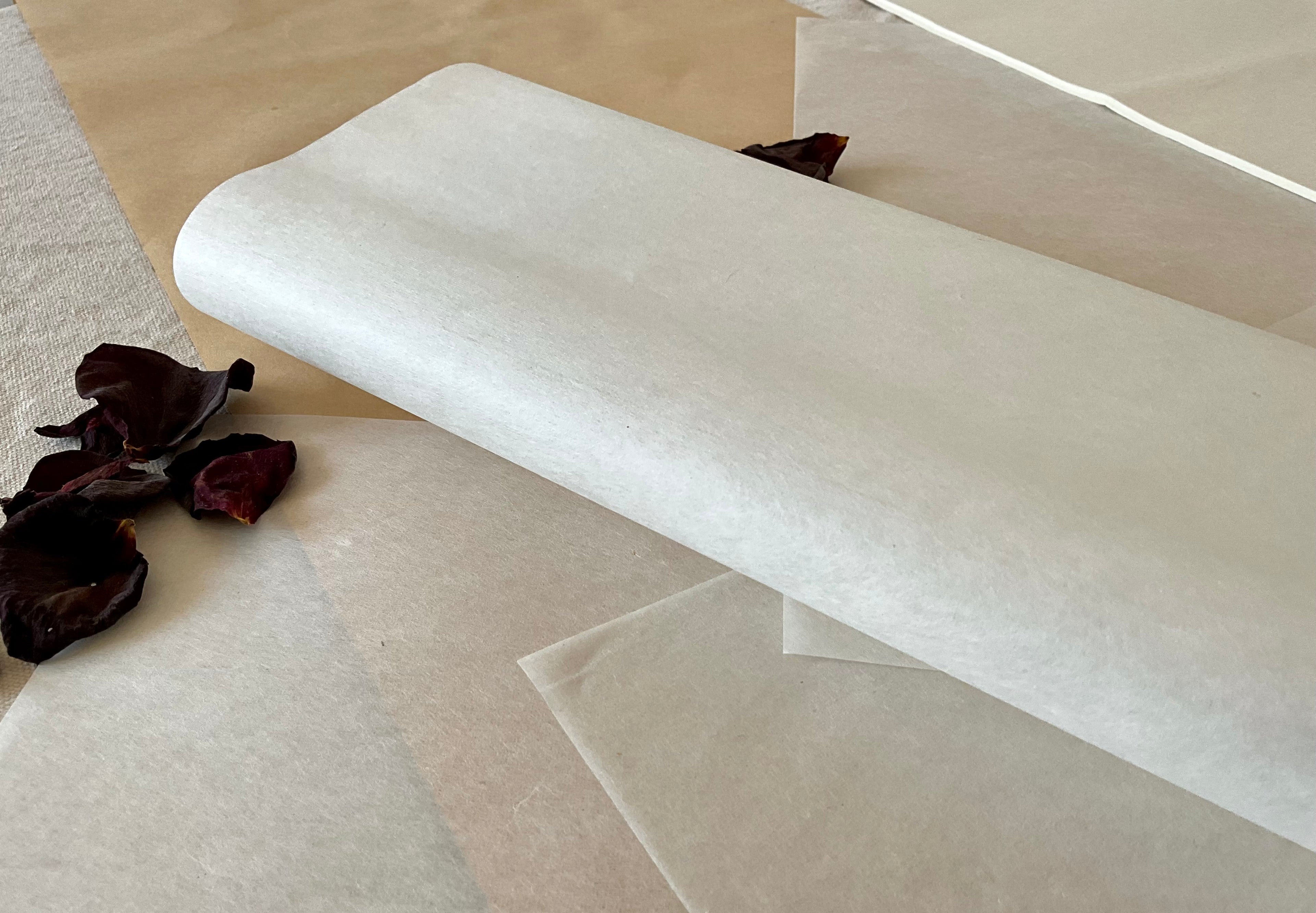 A loose roll of thin white Japanese hanshi paper resting at an angle on layered washi and kraft paper, dried dark rose petals scattered to the left, showing the translucent quality and soft texture of the sheet