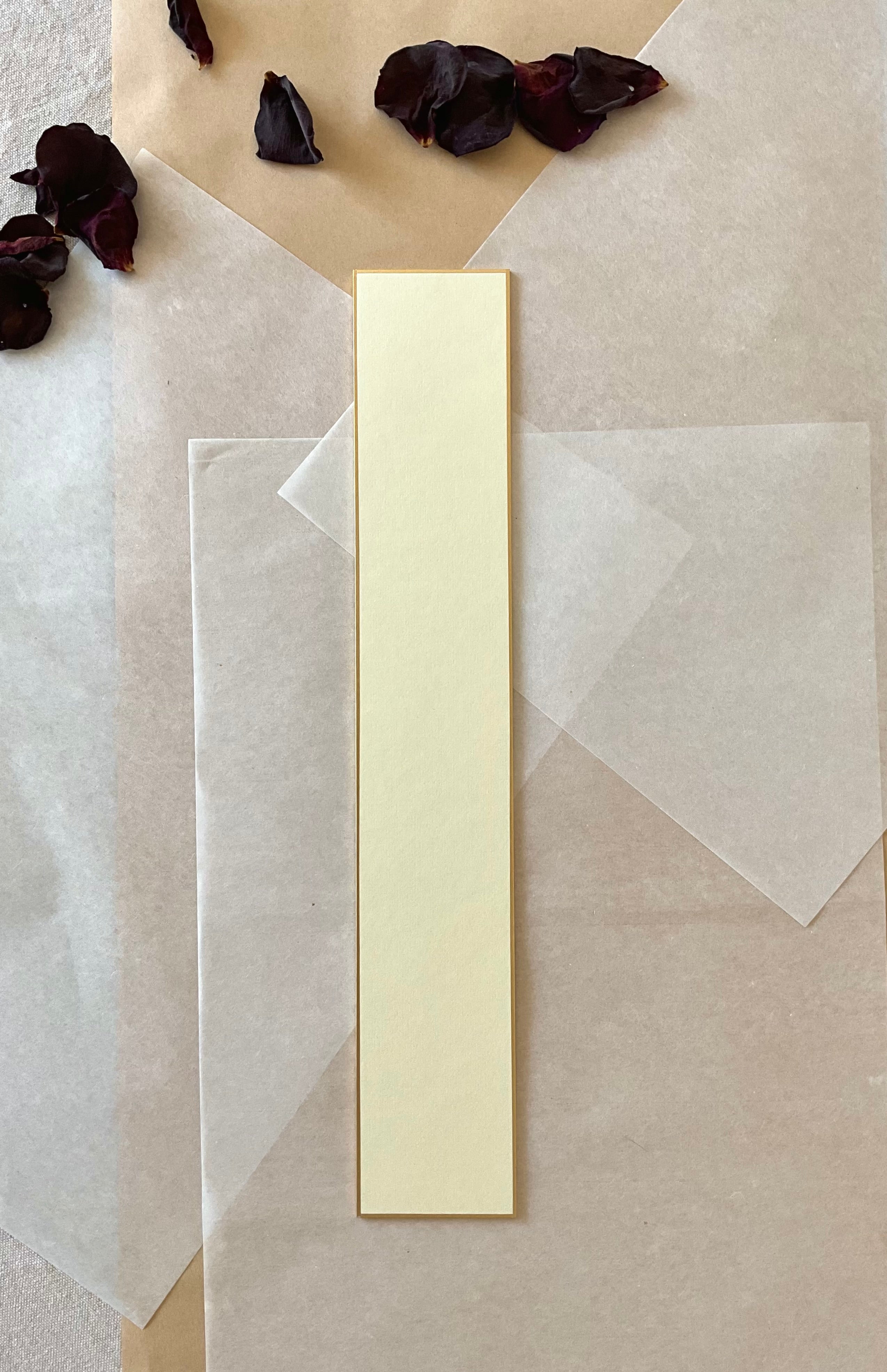 Single Kyukyodo tanzaku strip in cream with speckled texture and gold border, centered on layered washi and kraft paper with scattered dried rose petals, top-down view