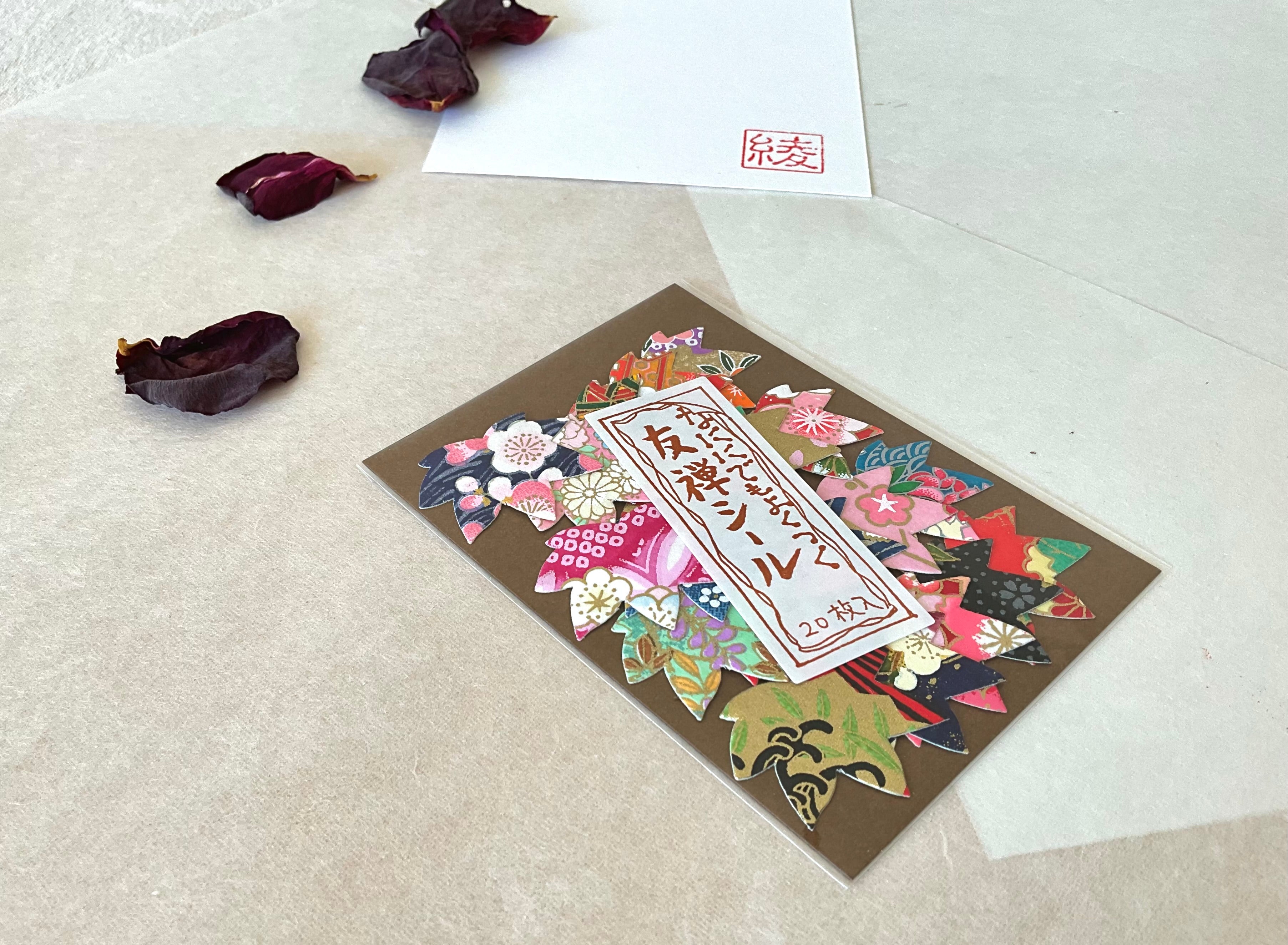 Morita Washi yuzen sticker pack at a slight angle on washi paper, showing flower and petal shaped sticker cuts in layered reds, pinks, dark navy and green chiyogami patterns, with dried rose petals in background