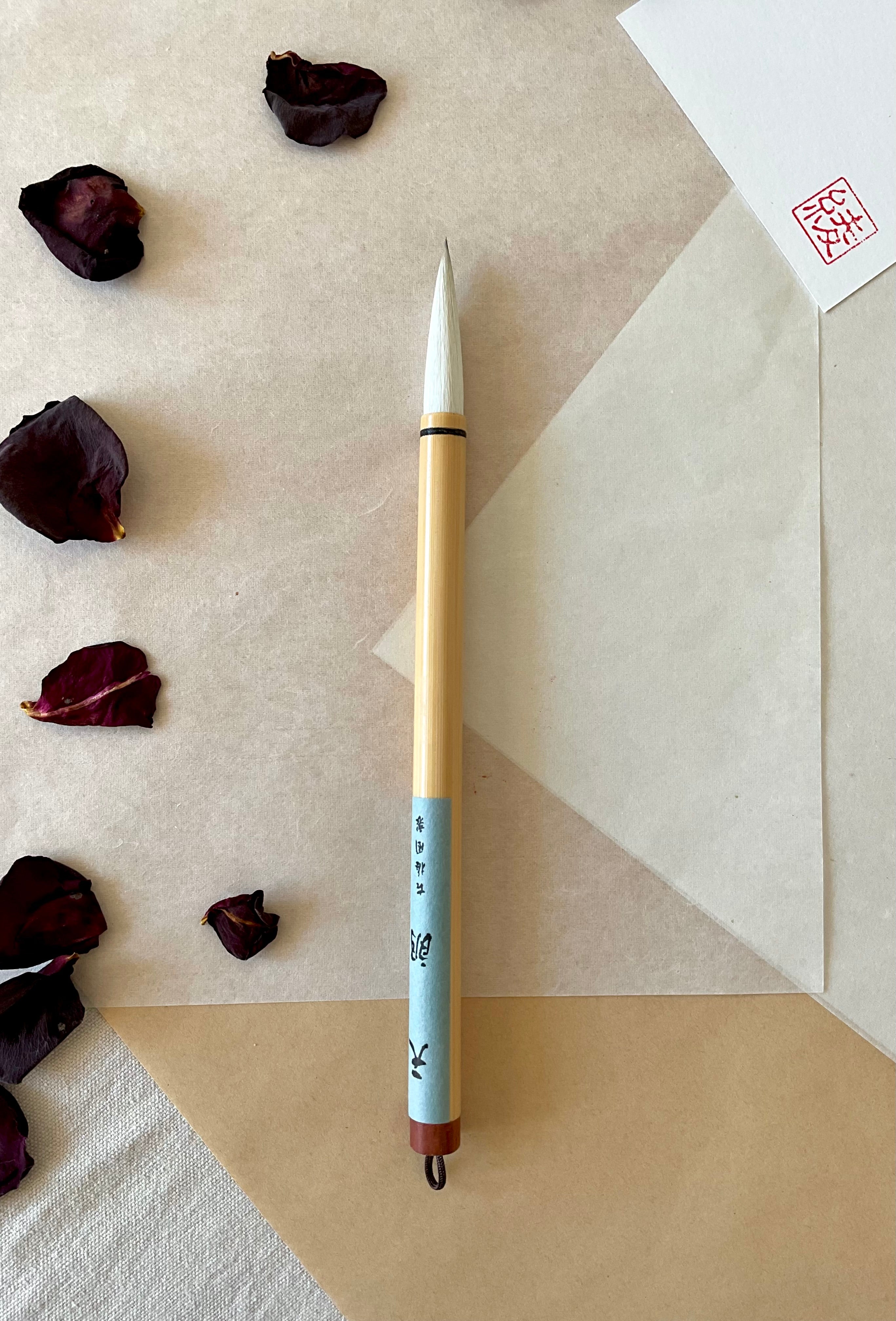 Kobaien large calligraphy brush with pale soft bristle and bamboo handle wrapped in light blue paper, hanging loop at the base, resting on layered washi paper with dried rose petals and a red hanko stamp card